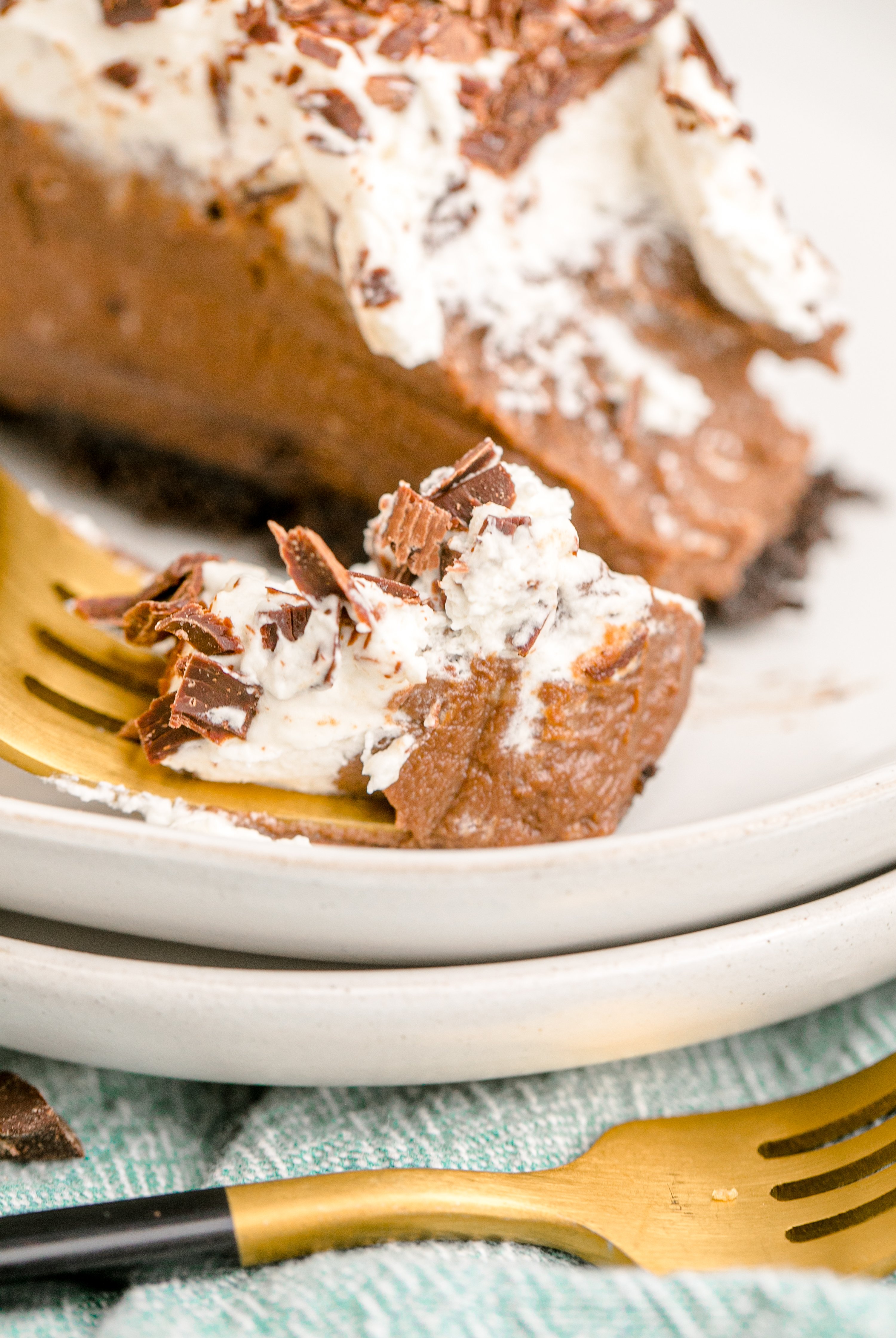 Chocolate Cream Pie topped with whipped cream and chocolate shavings on a white plate with a gold fork bite shot