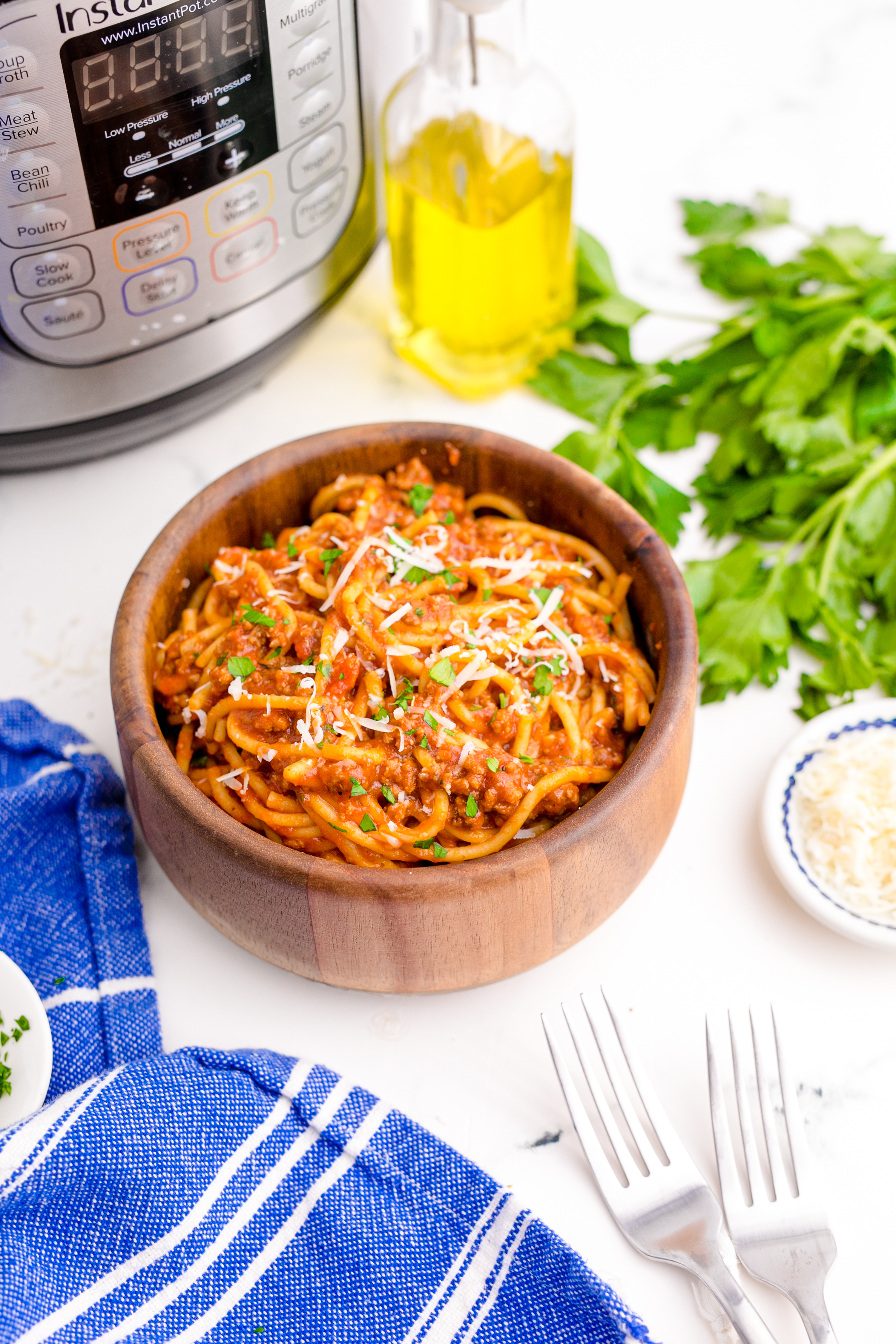 Instant Pot Spaghetti in a wooden bowl with a blue napkin next to the bowl , a fork , the instant pot in the background and a small bowl of parmesan cheese on the side