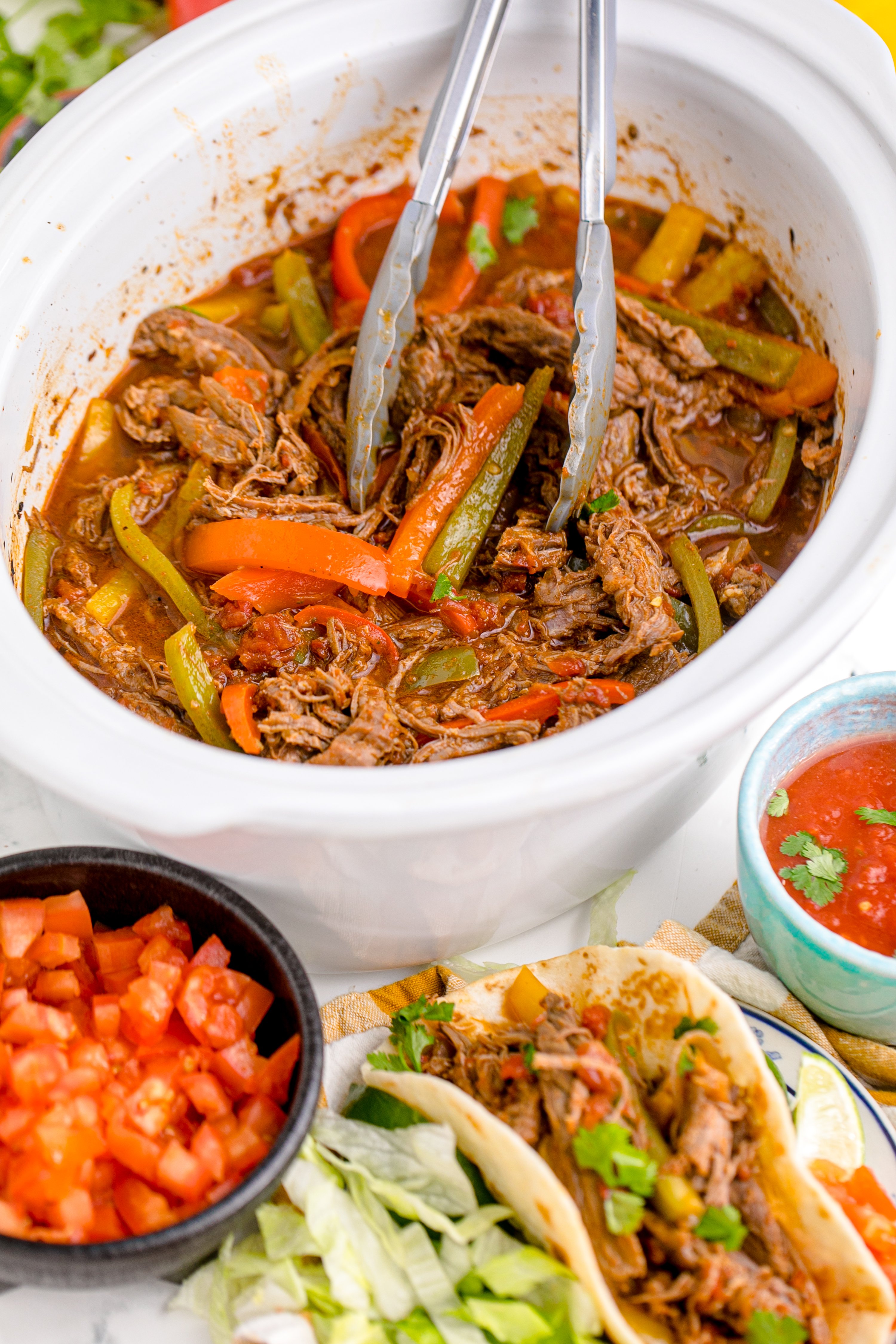 Steak Fajitas in a crock pot with tongs lifting the steak, with a bowl of salasa next to it
