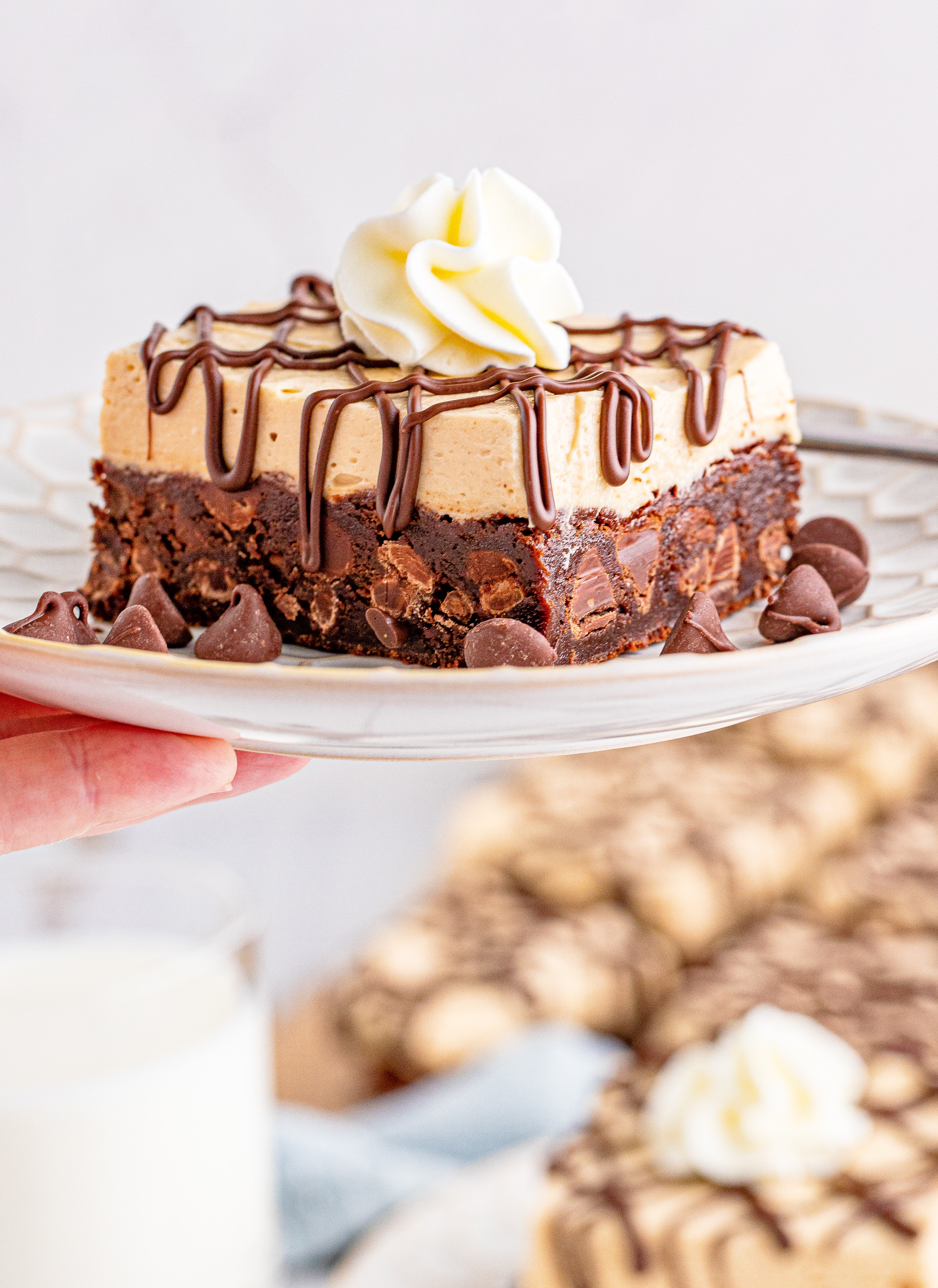 a hand holding up a plate with Peanut Butter Brownies on a white place drizzeled with chocolate and topped with whipped cream