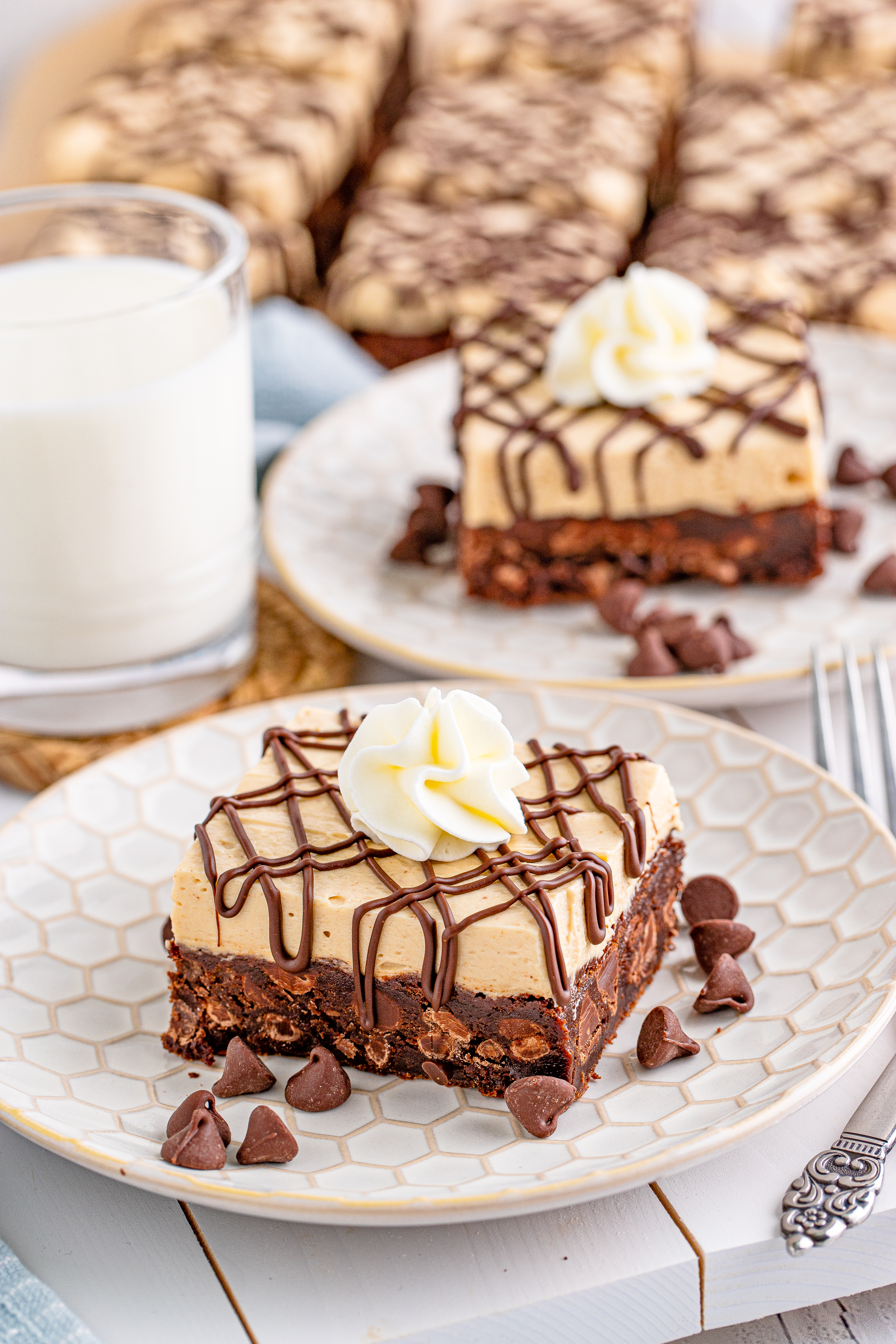 Peanut Butter Brownies on a white place drizzled with chocolate and topped with whipped cream with a cup of milk beside it and chocolate morsels on the plate
