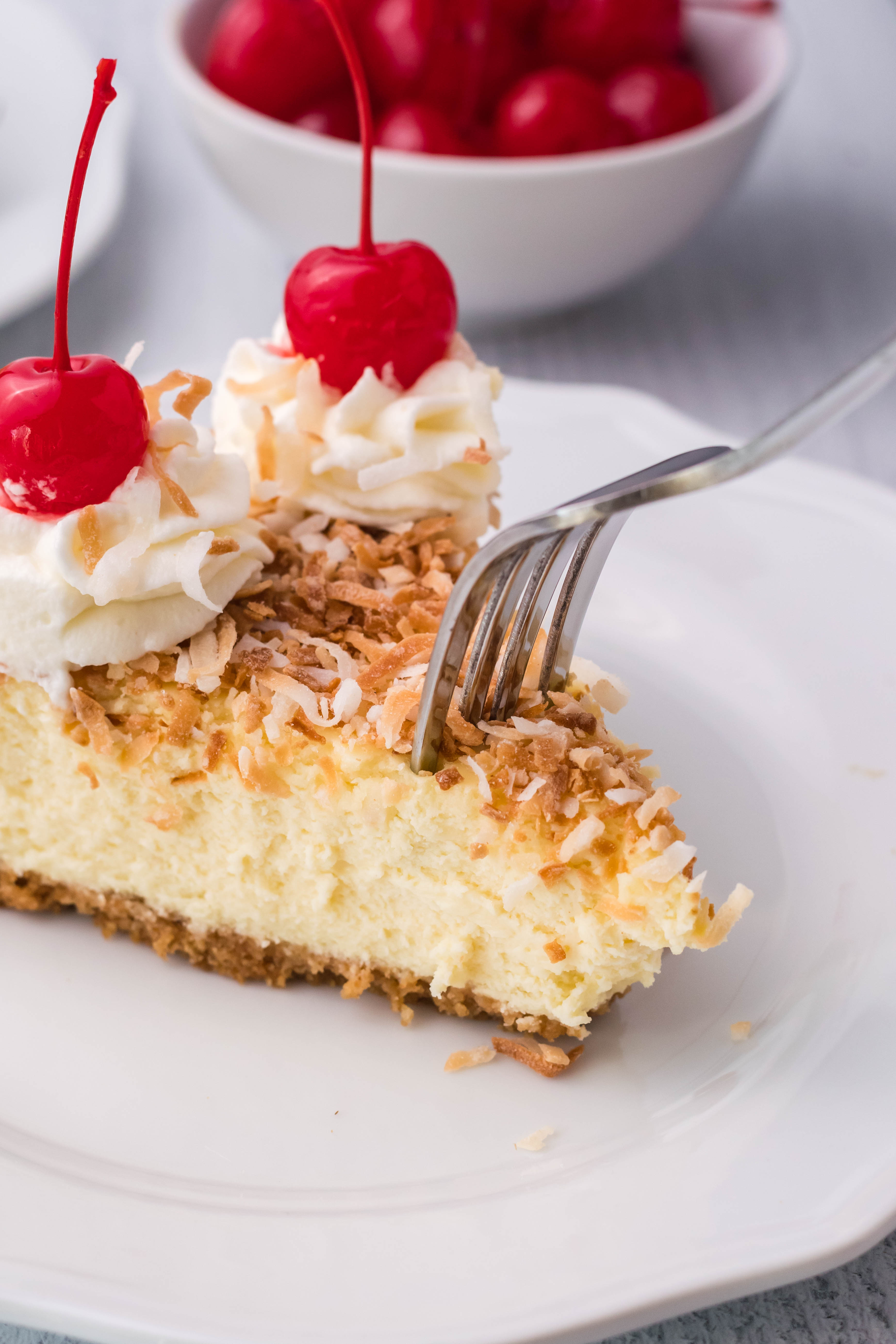 Coconut Cheesecake on a white plate topped with cherries and whipped cream with a fork slicing into it
