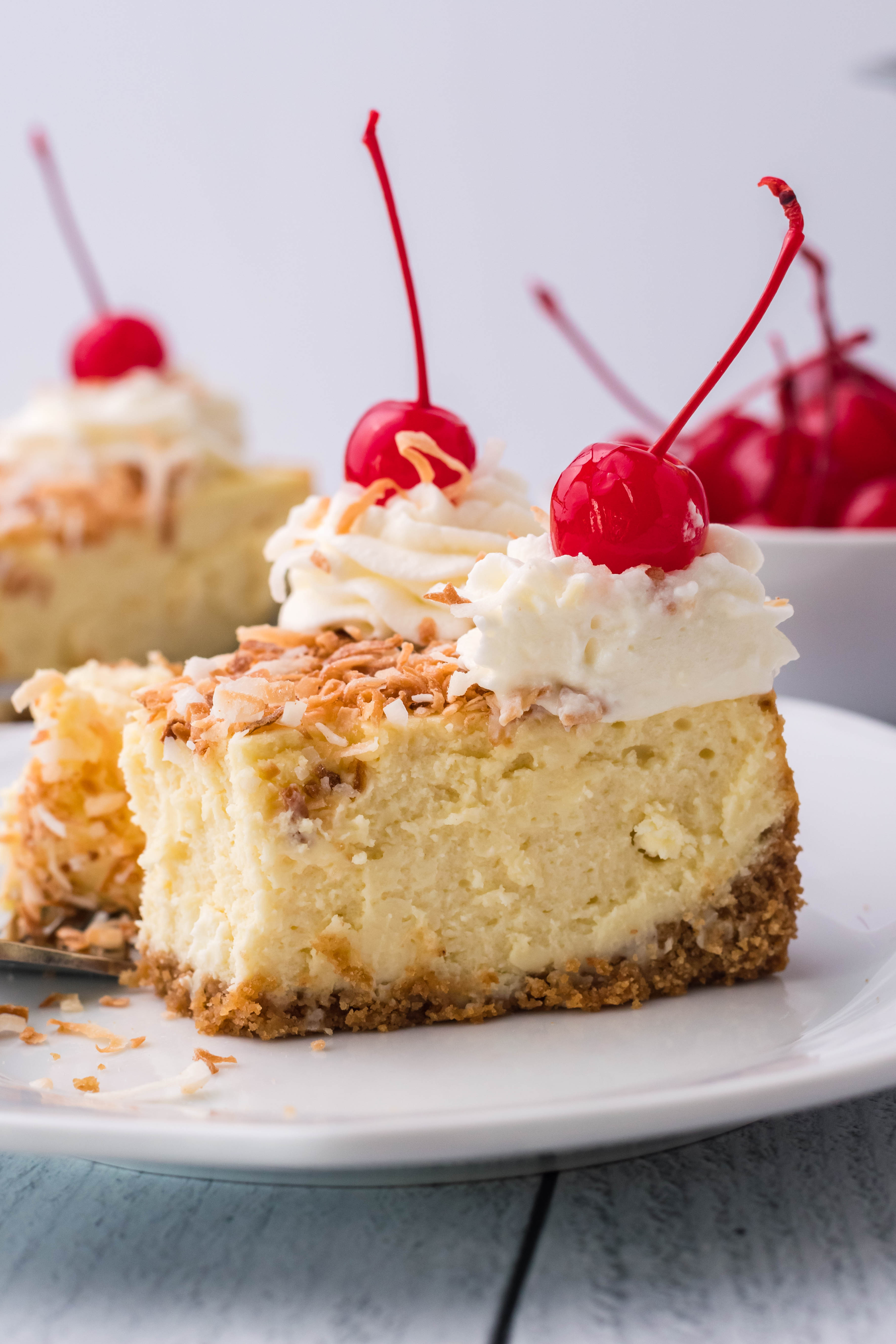 Coconut Cheesecake on a white plate topped with cherries and whipped cream side shot