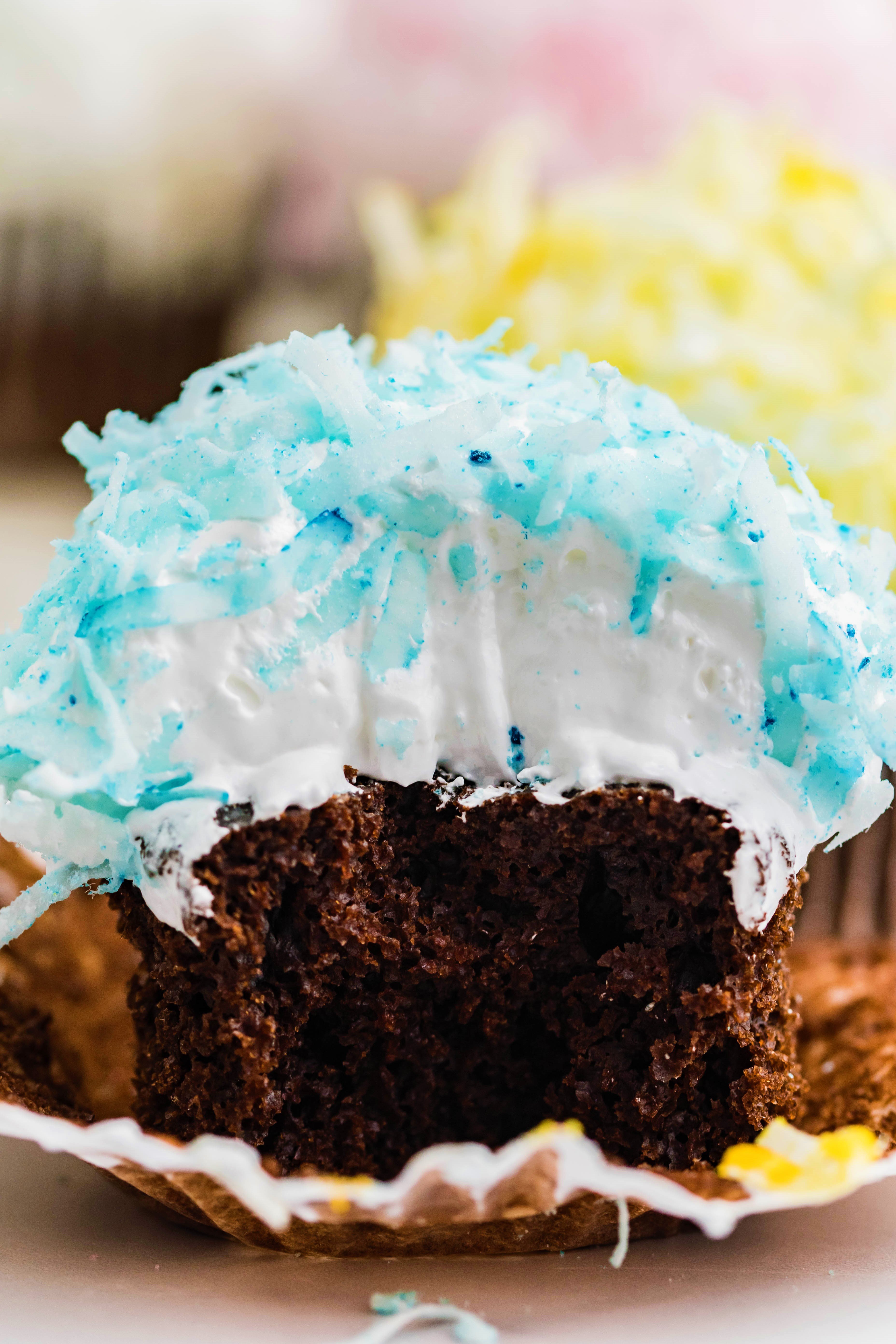 chocolate cupcake with blue coconut shavings, marshmallow frosting with a bite taken out of it
