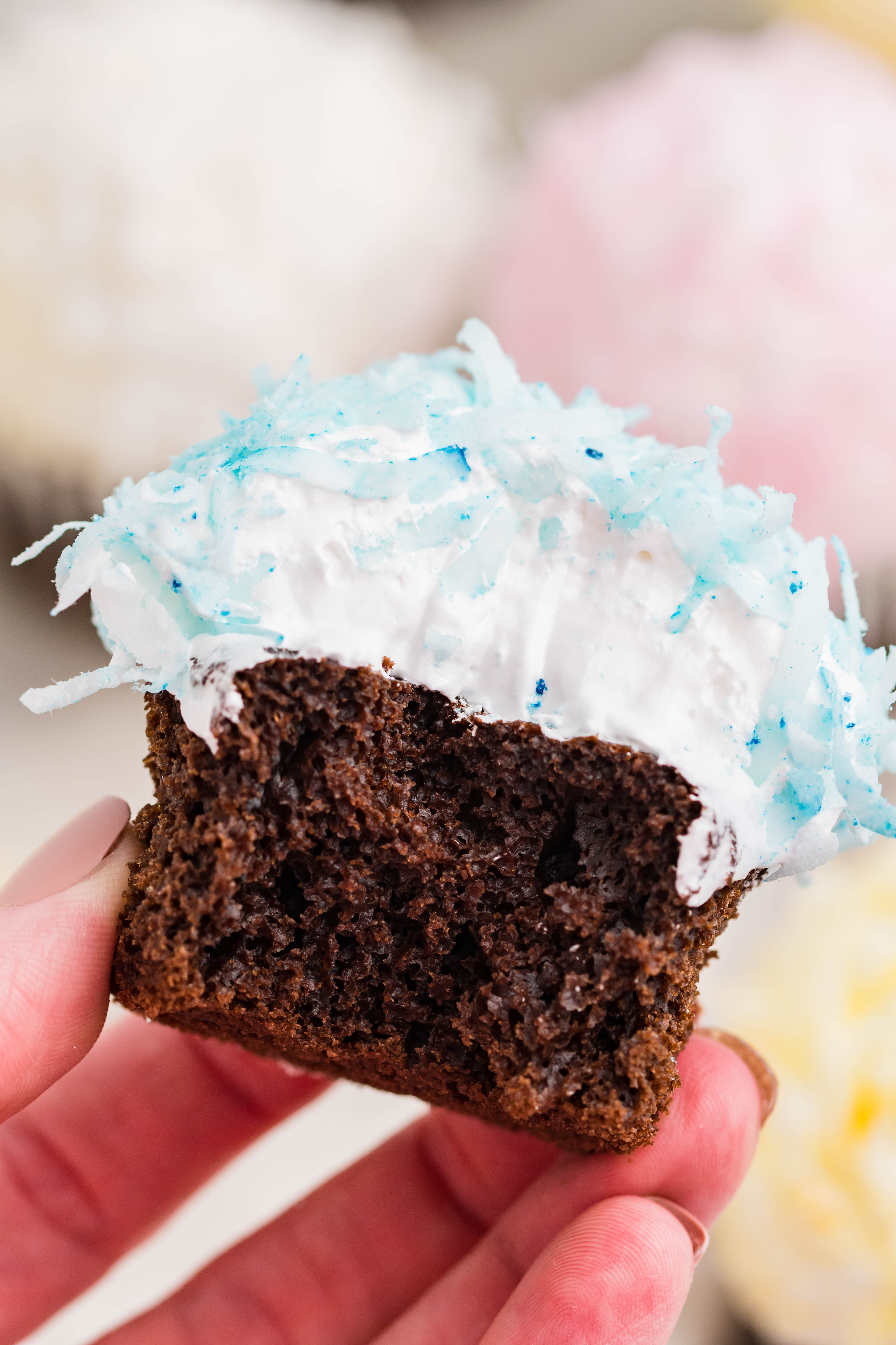 chocolate cupcake with blue coconut shavings, marshmallow frosting being held up with a bite taken out of it