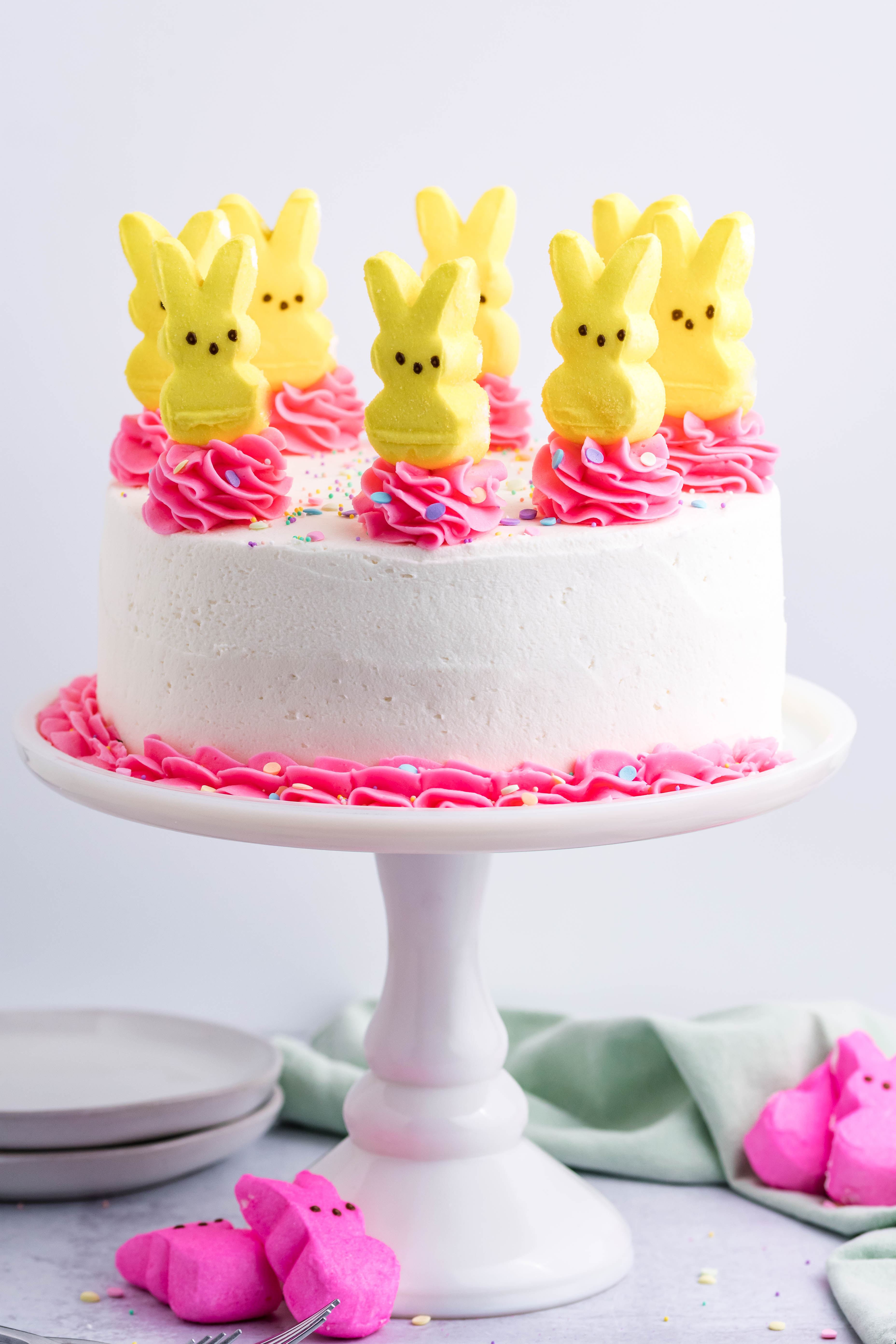 Pink Vanilla layered cake with white frosting and topped with yellow peeps for easter on a white cake stand