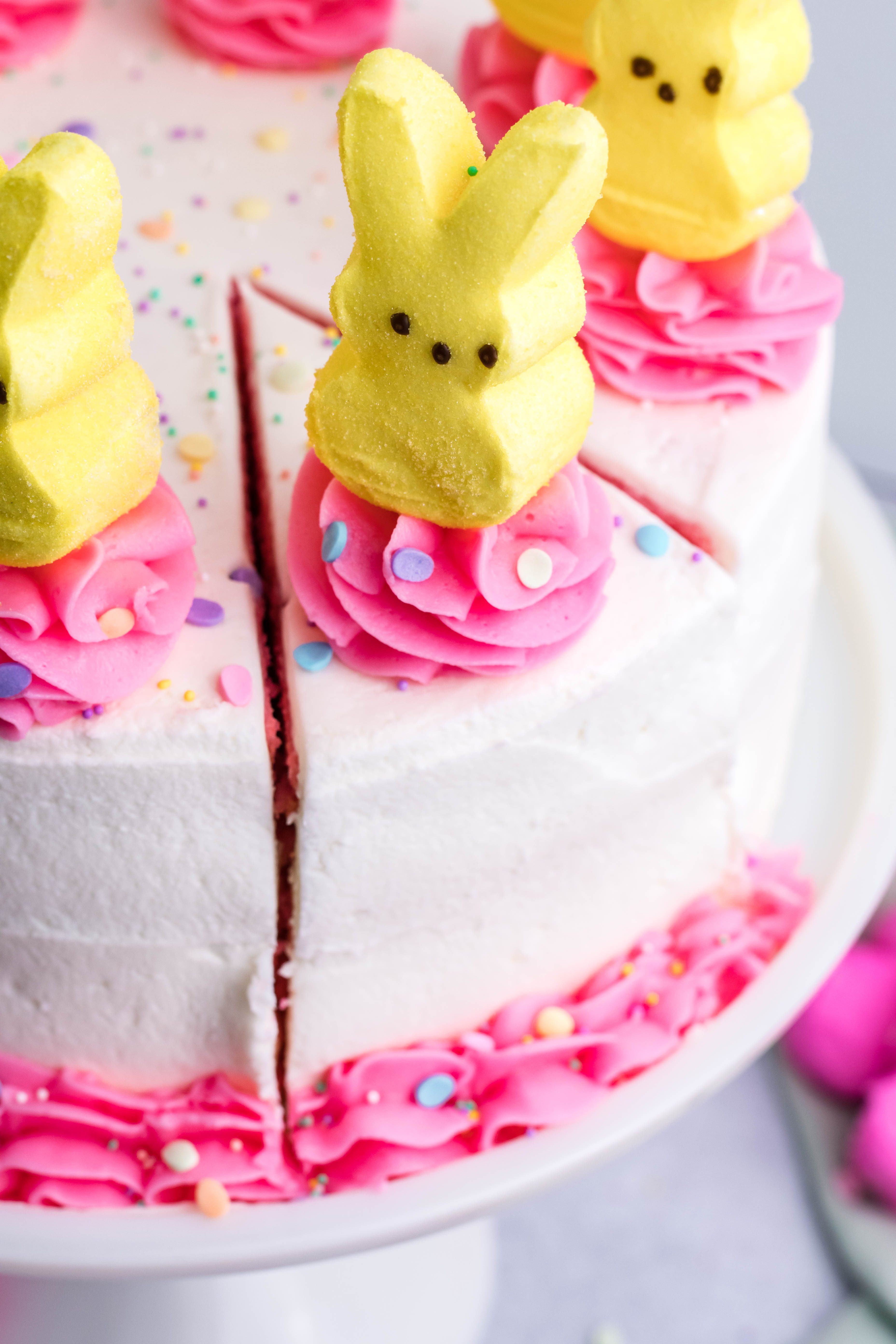 Pink Vanilla layered cake with white frosting and topped with yellow peeps for easter on a white cake stand overhead shot