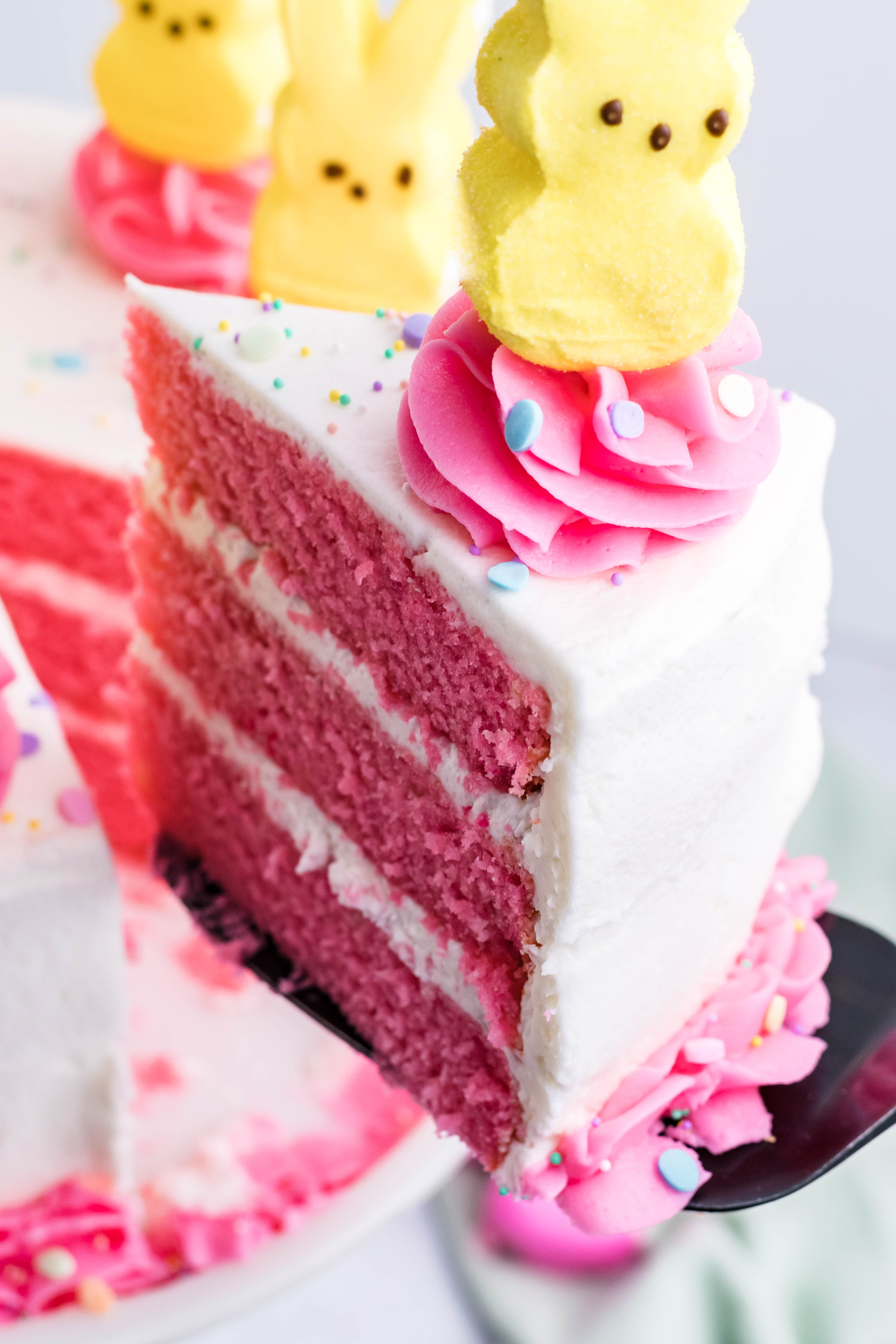 Pink Vanilla layered cake with white frosting and topped with yellow peeps for easter on a white cake stand being lifted up by a black cake cutter