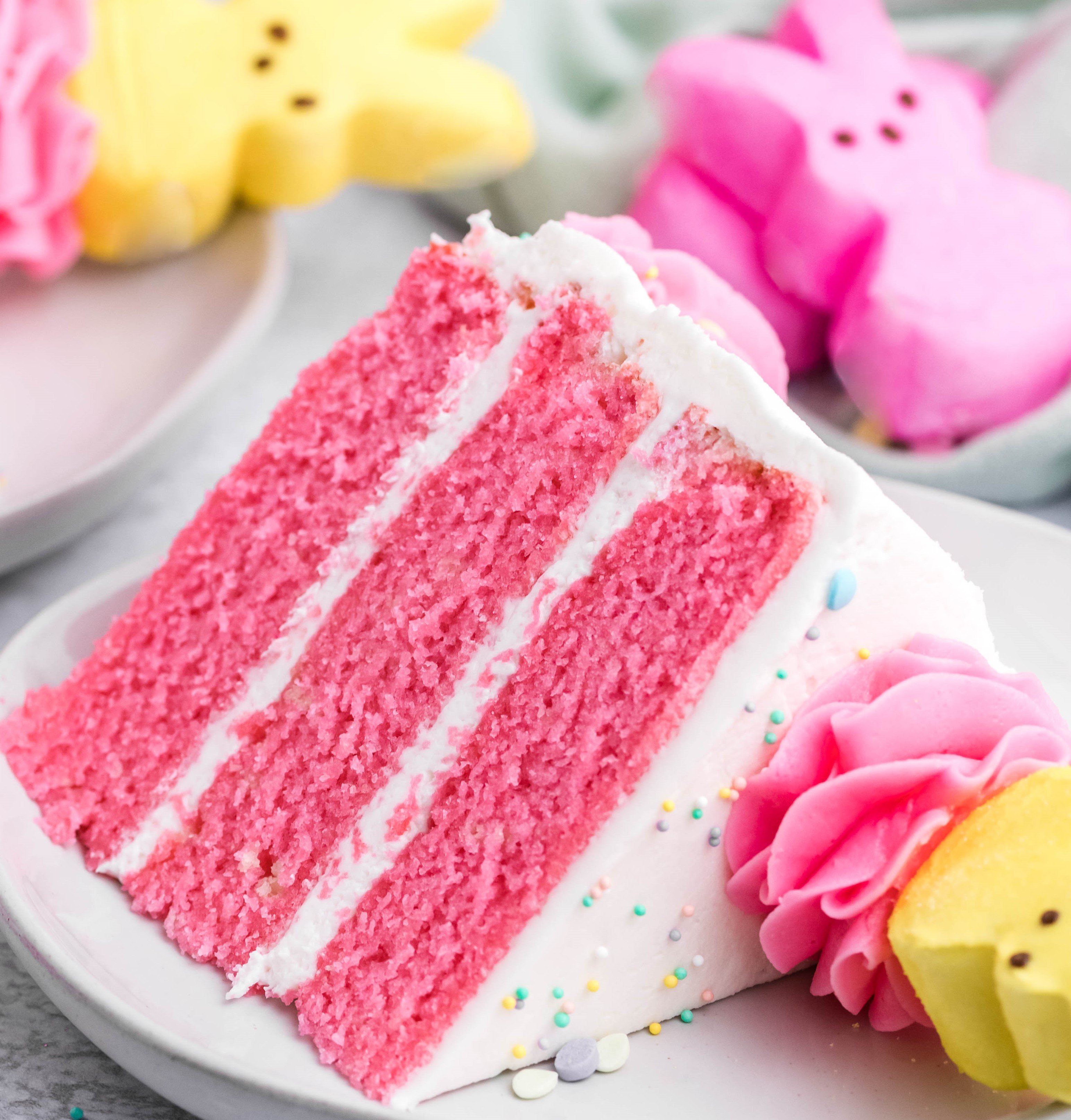 Pink Vanilla layered cake with white frosting and topped with yellow peeps for easter on a white plate with a bit gone