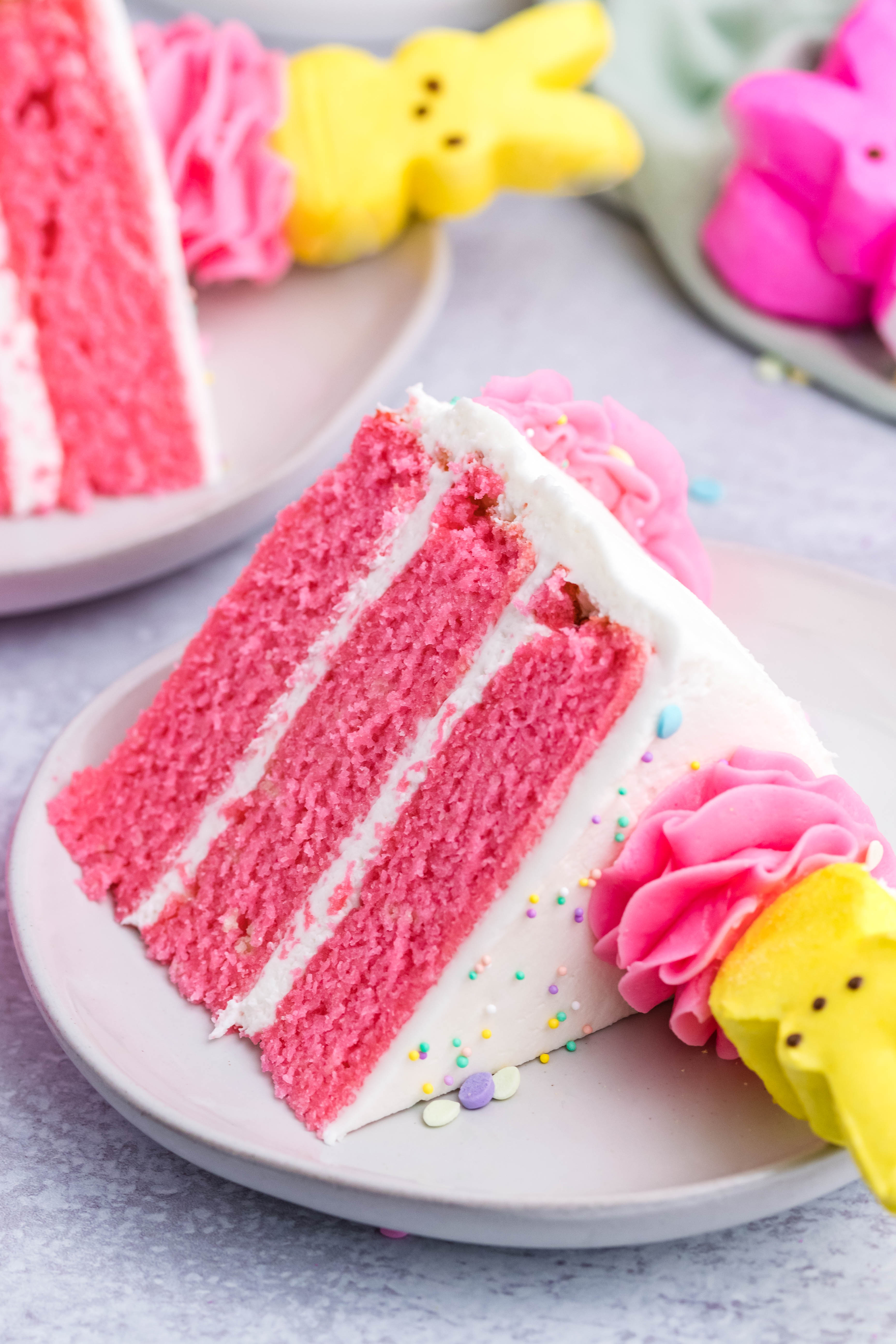 Pink Vanilla layered cake with white frosting and topped with yellow peeps for easter on a white plate with two other slices next to it