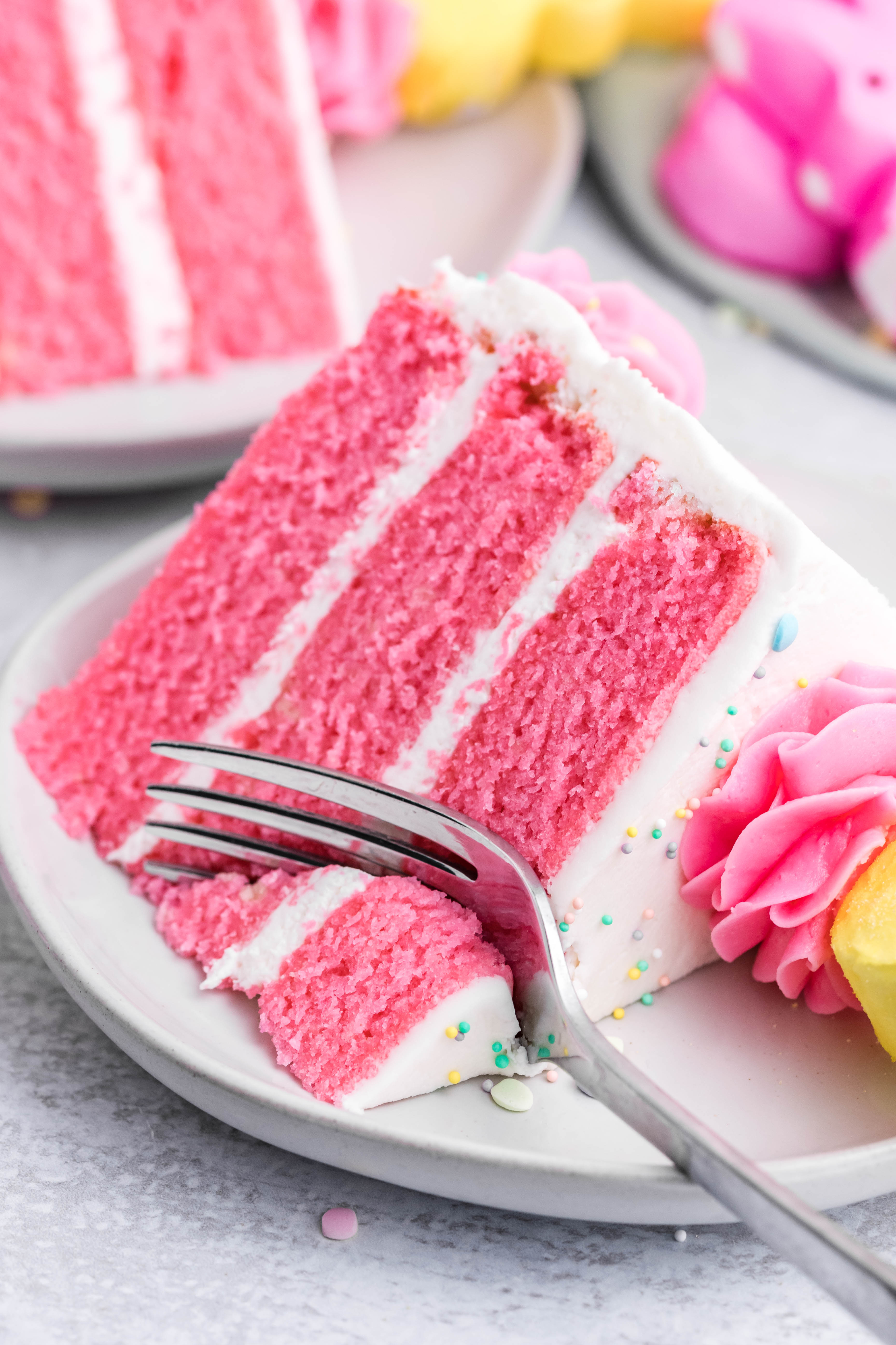 Pink Vanilla layered cake with white frosting and topped with yellow peeps for easter on a white plate with a fork slicing into the cake