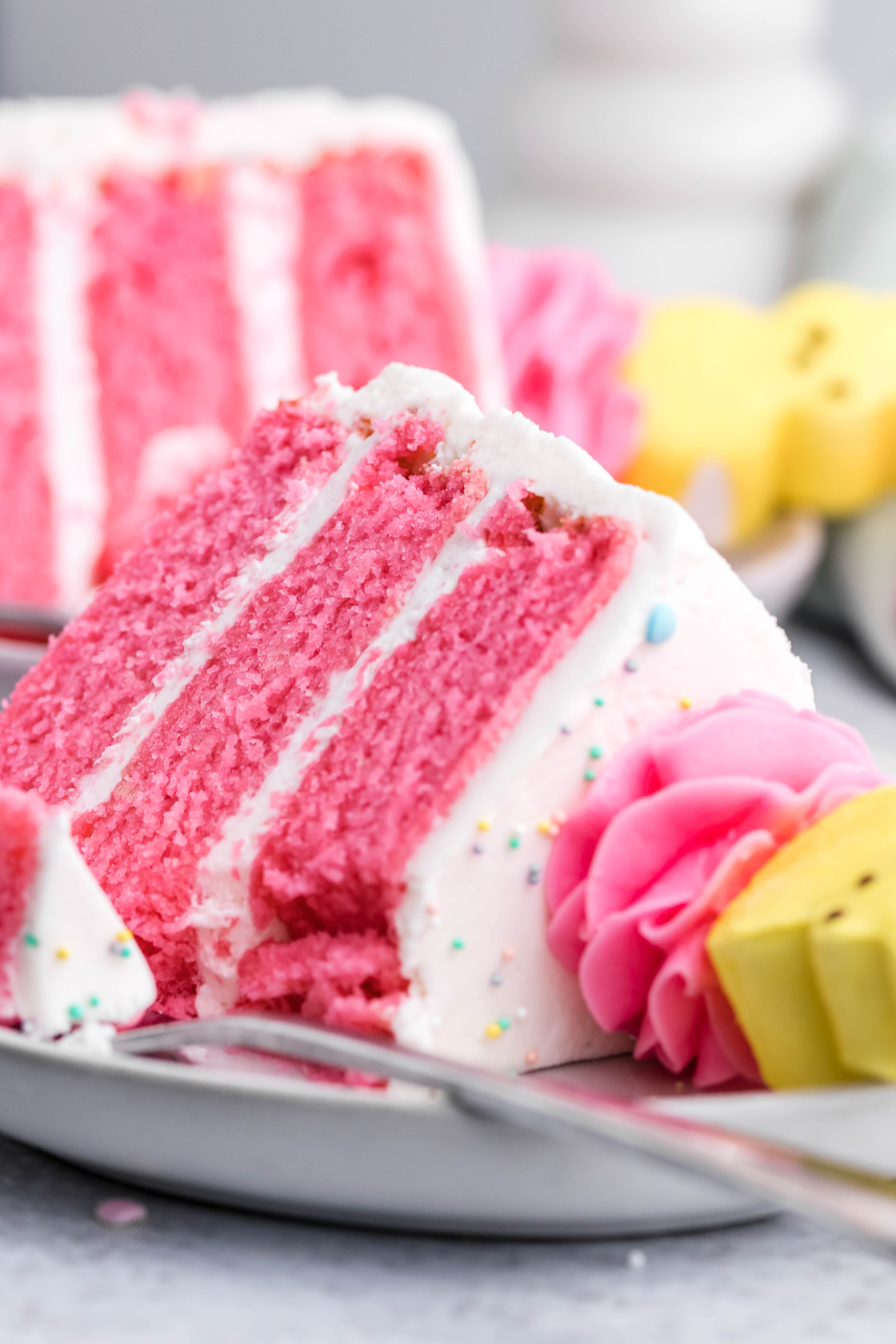 Pink Vanilla layered cake with white frosting and topped with yellow peeps for easter on a white plate with a bit gone