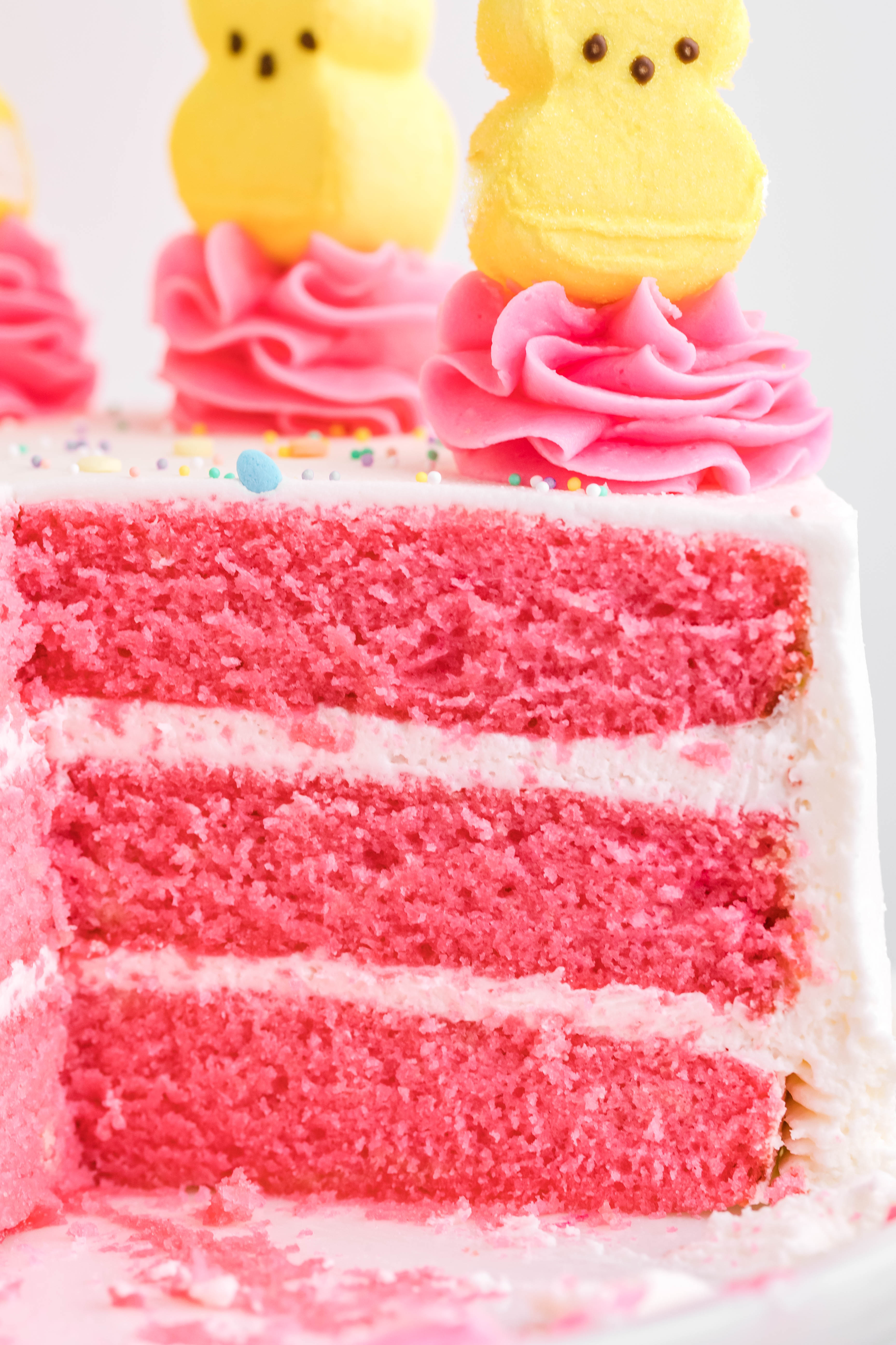 Pink Vanilla layered cake with white frosting and topped with yellow peeps for easter on a white cake stand from the side to show a close up of all the hot pink layers
