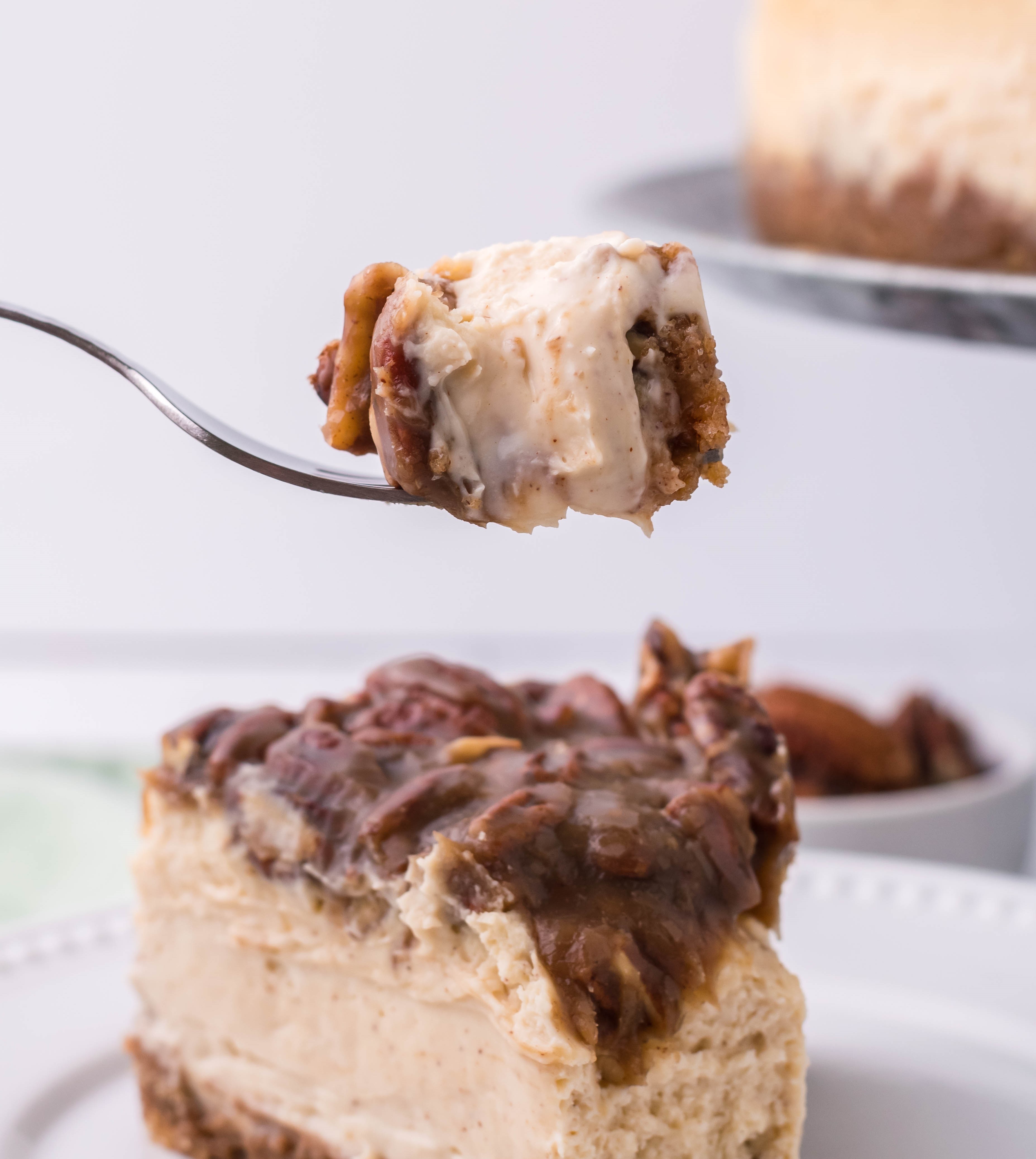 pecan pie Cheesecake slice on a white plate with a bite being lifted up on a fork