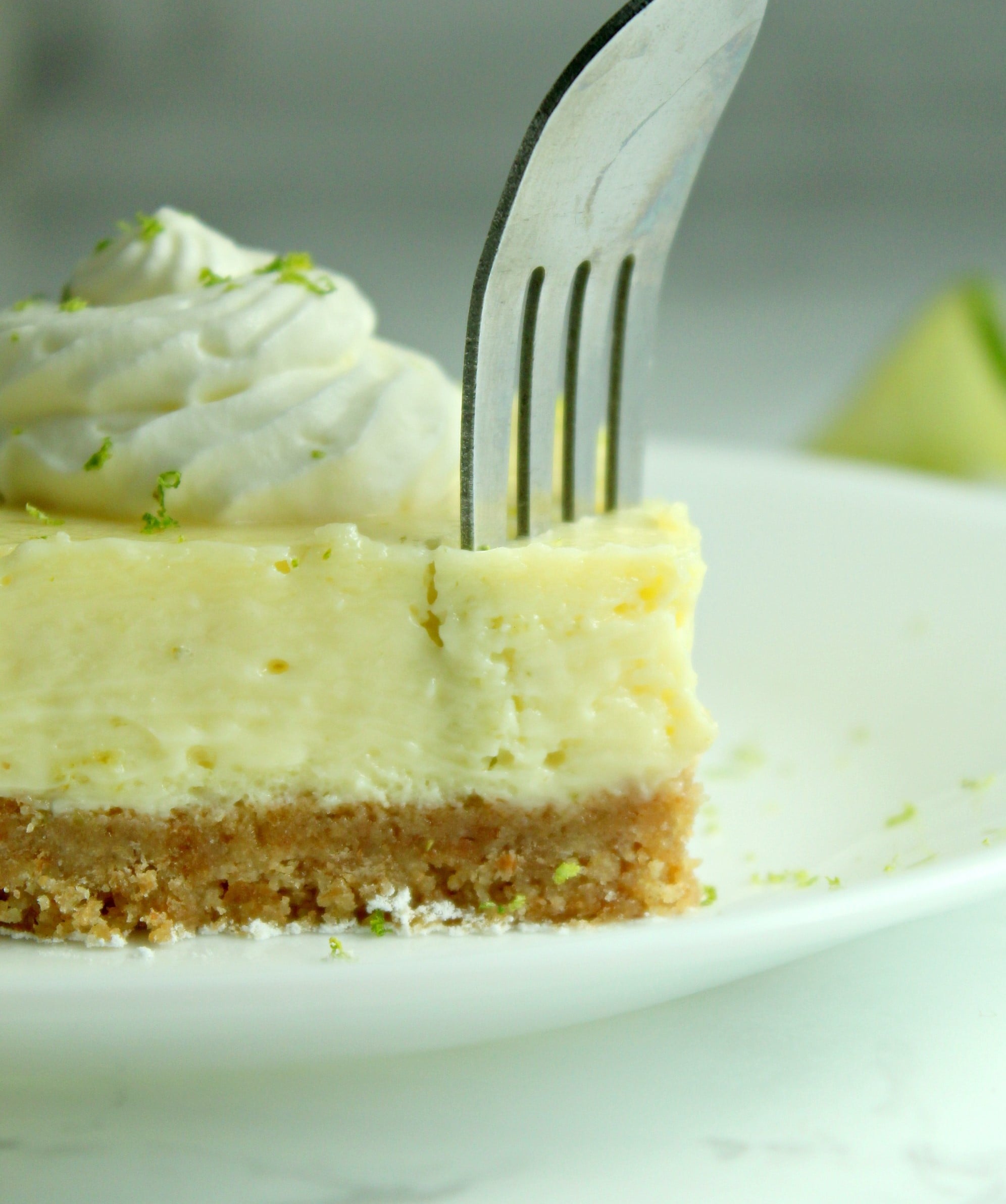 Key Lime Bars with lime whipped cream topped with lime zest on a white plate with a fork cutting into it