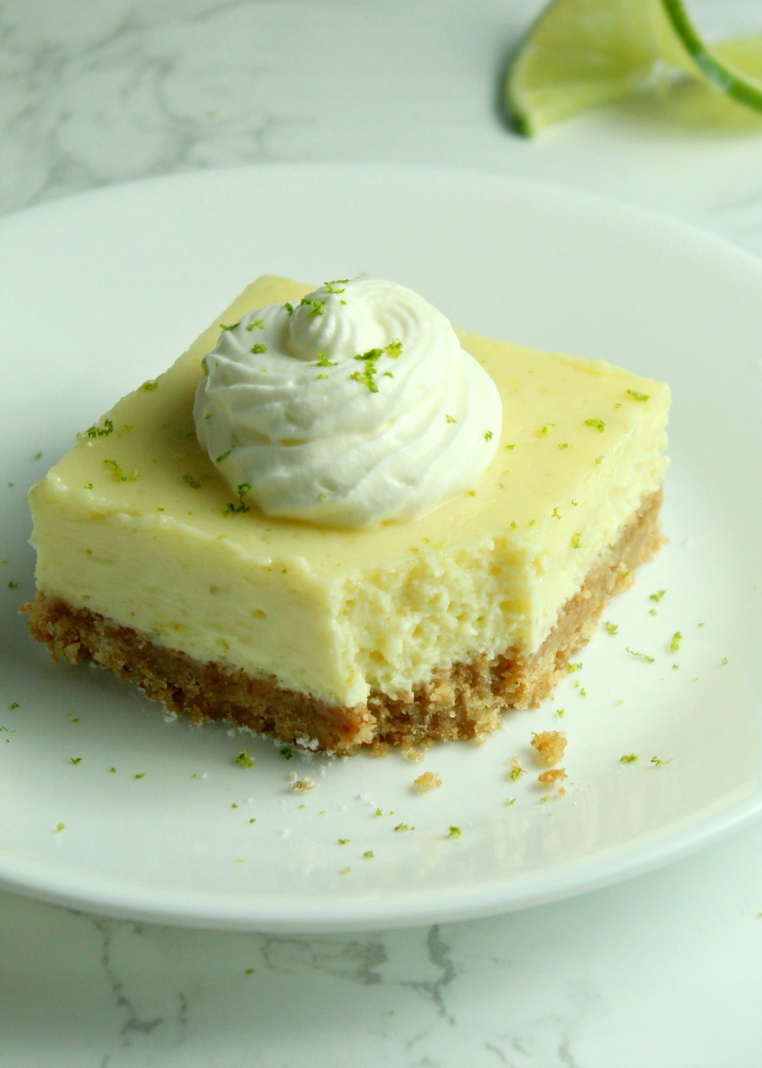 Key Lime Bars with lime whipped cream topped with lime zest on a white plate with a bite gone