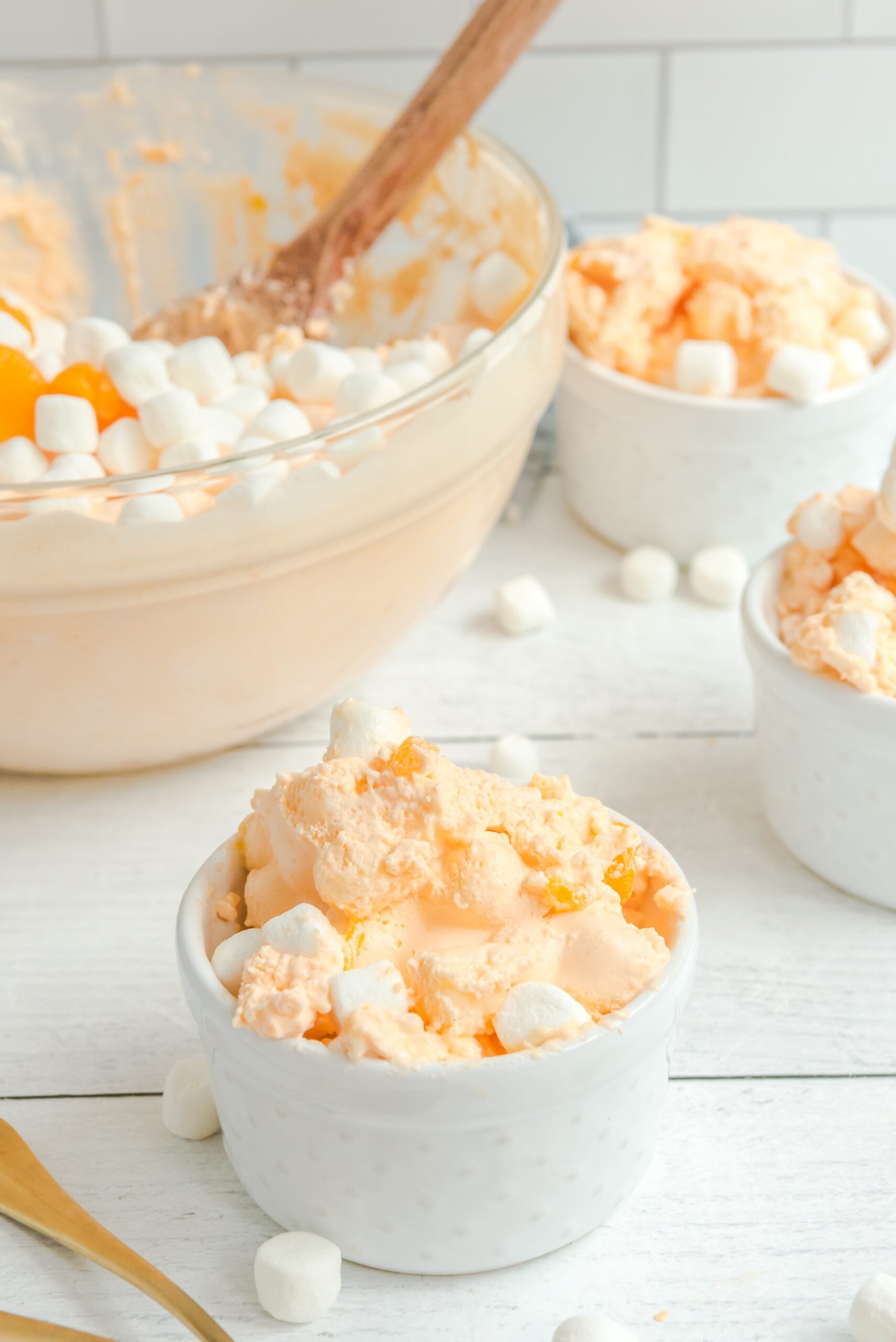 Orange Creamsicle Salad in a white bowl topped with marshmallows and mandarin oranges with a huge glass bowl behind the three small bowls
