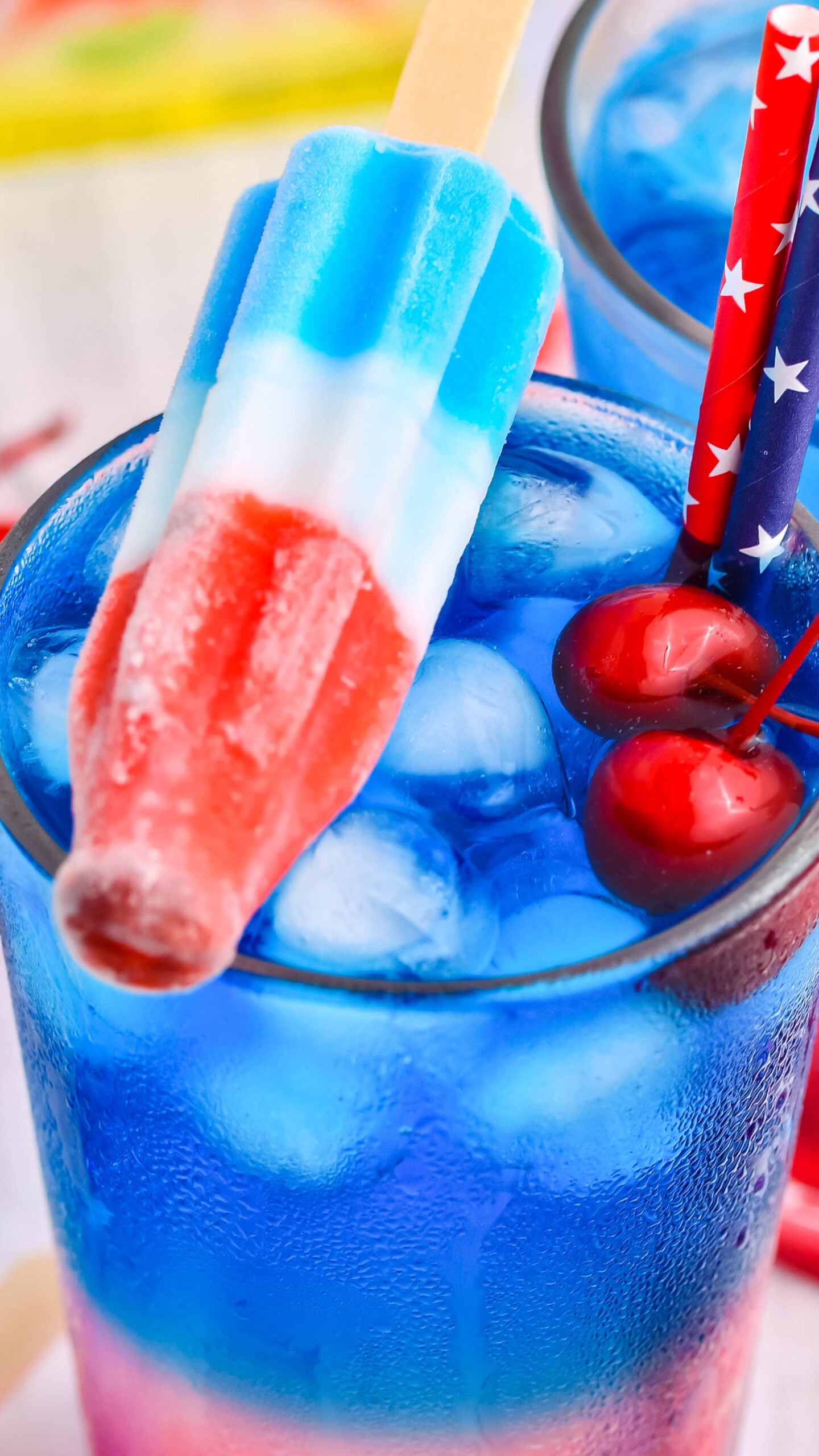 Bomb Pop Cocktail Drink in a clear glass with a blue & red straw with cherries floating up close