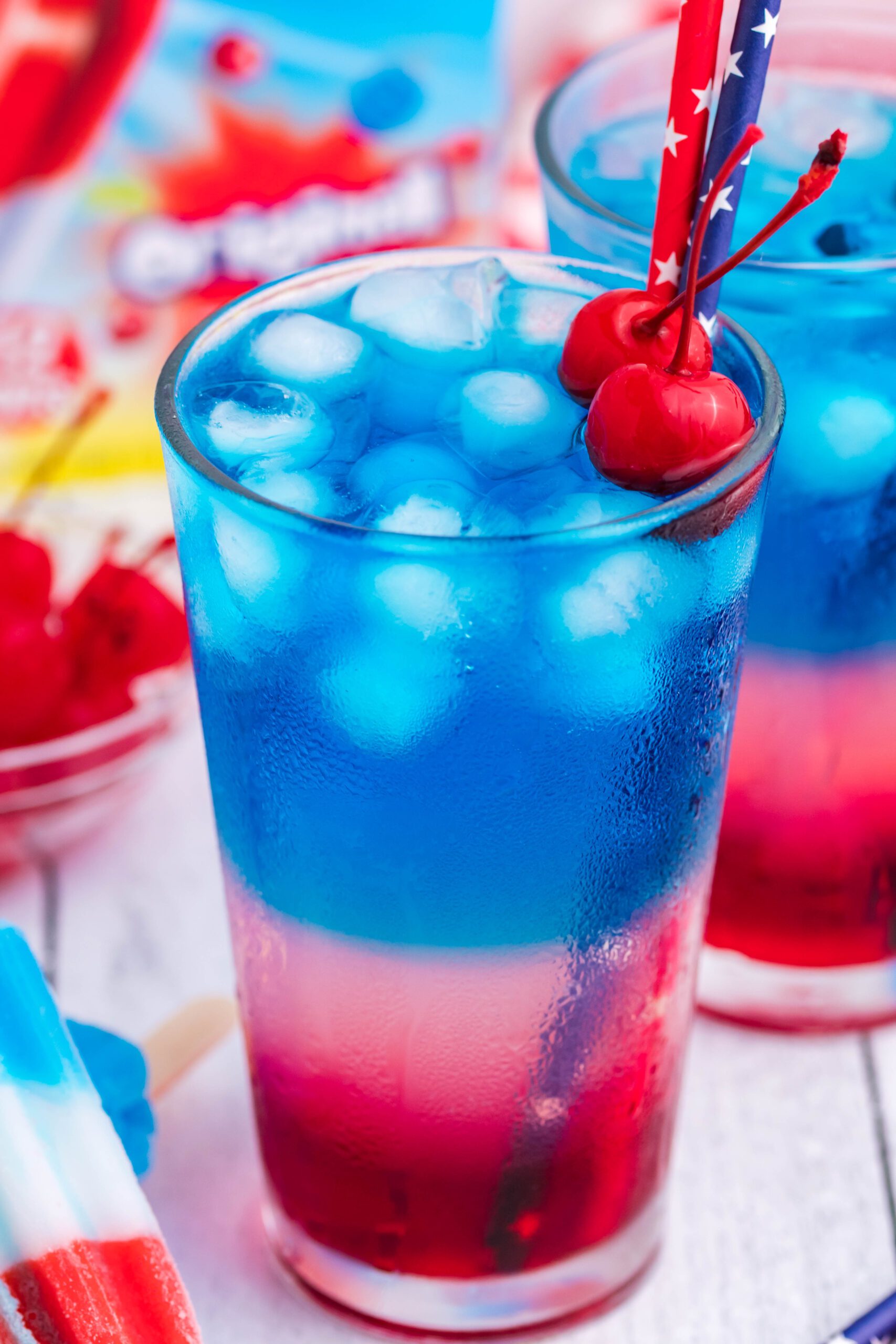 Bomb Pop Cocktail Drink in a clear glass with a blue & red straw with cherries floating