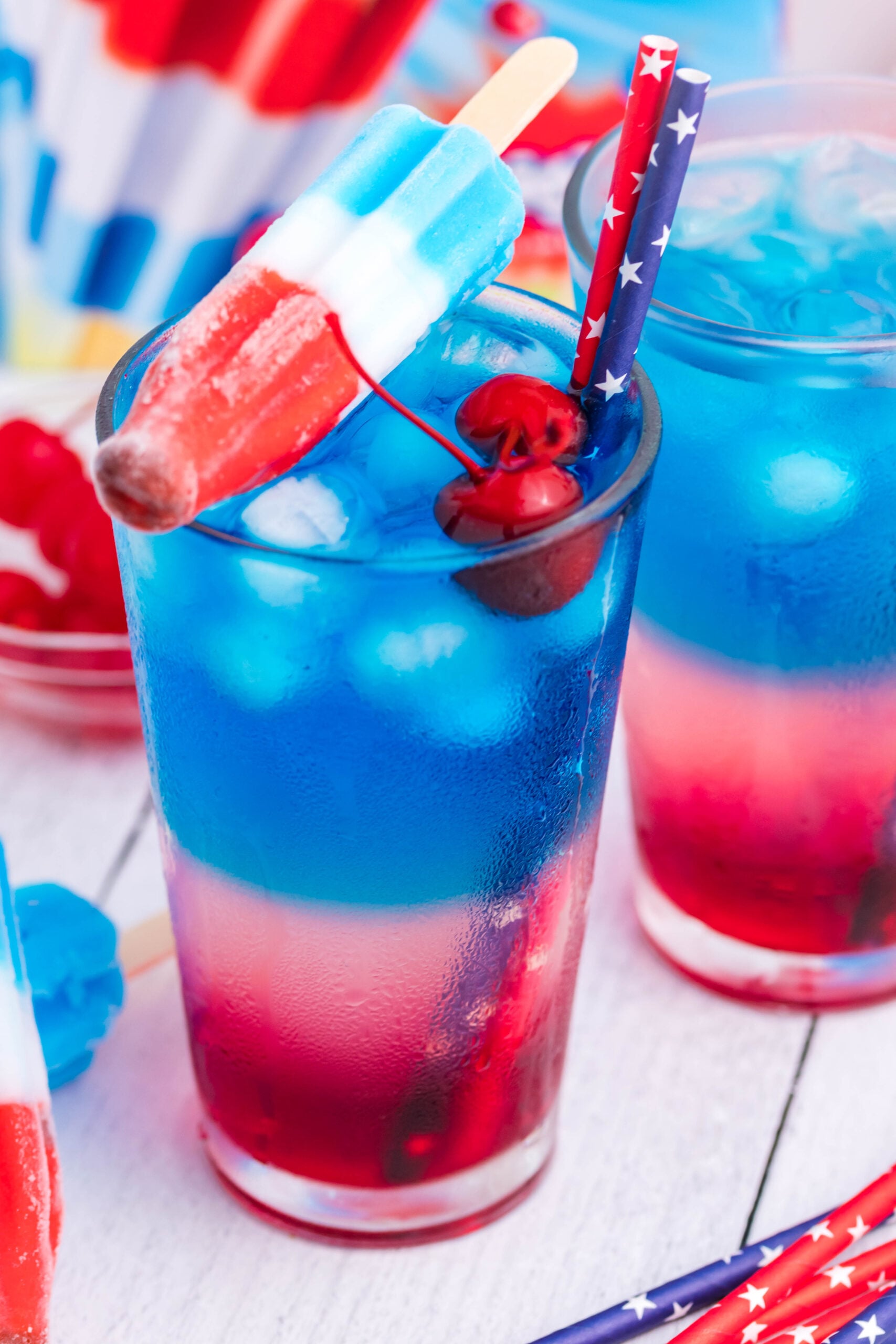 Bomb Pop Cocktail Drink in a clear glass with a blue & red straw with cherries floating and a ice cream sitting on top