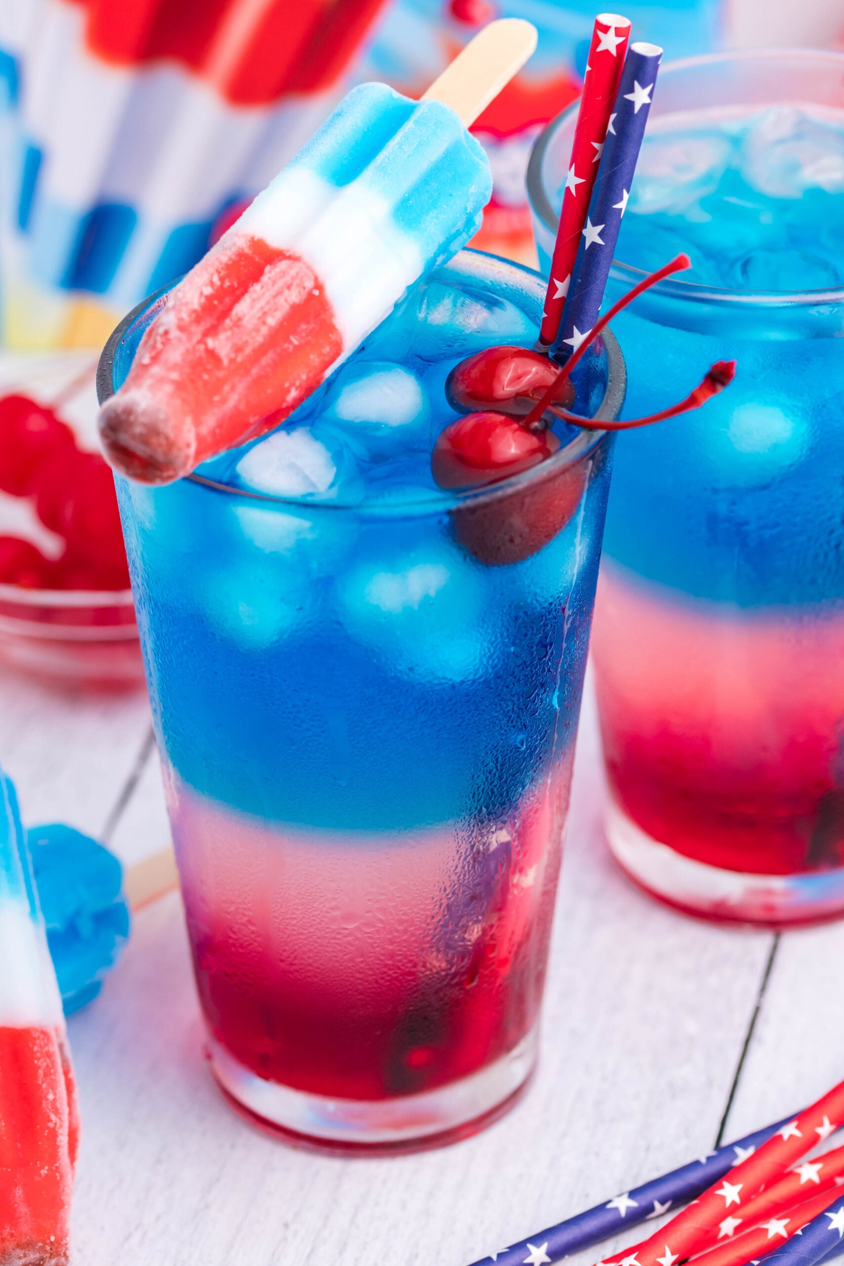 Bomb Pop Cocktail Drink in a clear glass with a blue & red straw with cherries floating