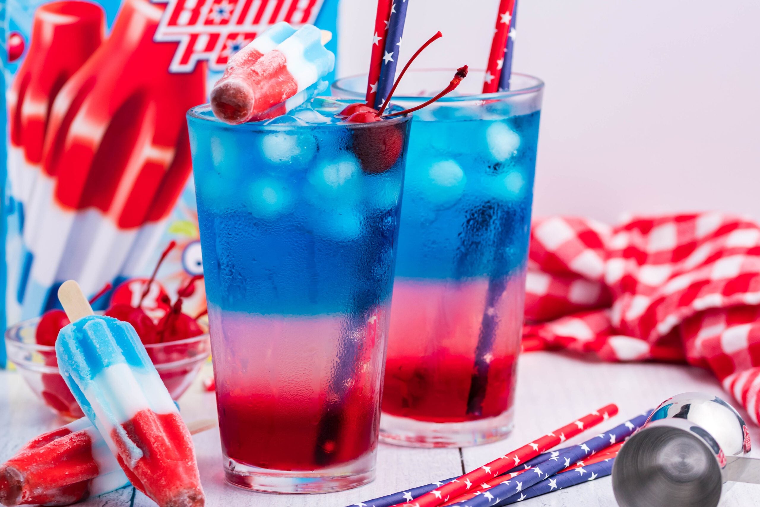 two Bomb Pop Cocktail Drink in a clear glass with a blue & red straw with cherries floating