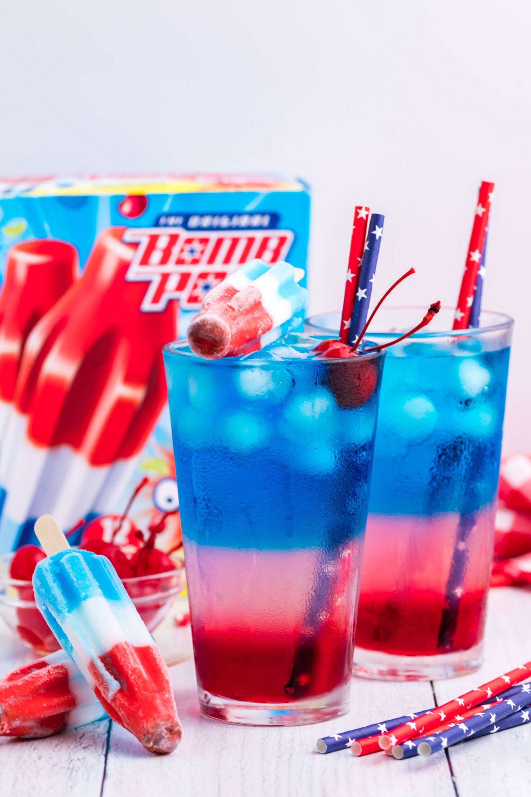 Bomb Pop Cocktail Drink in a clear glass with a blue & red straw with cherries floating far away shot