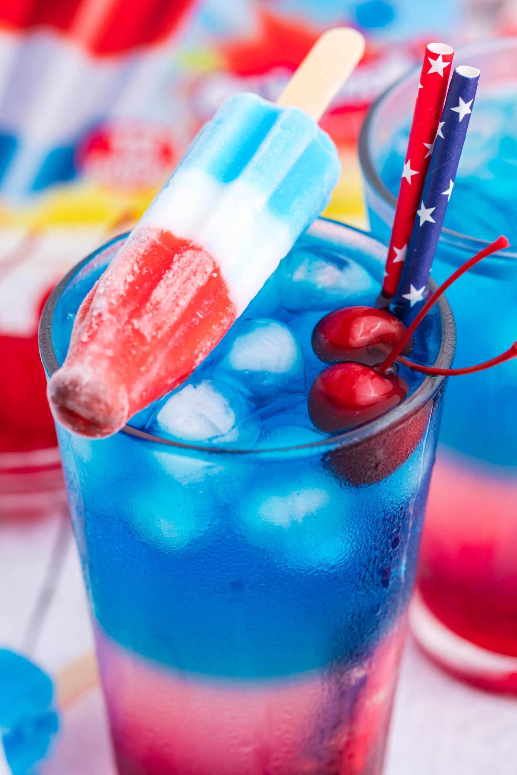 Bomb Pop Cocktail Drink in a clear glass with a blue & red straw with cherries floating