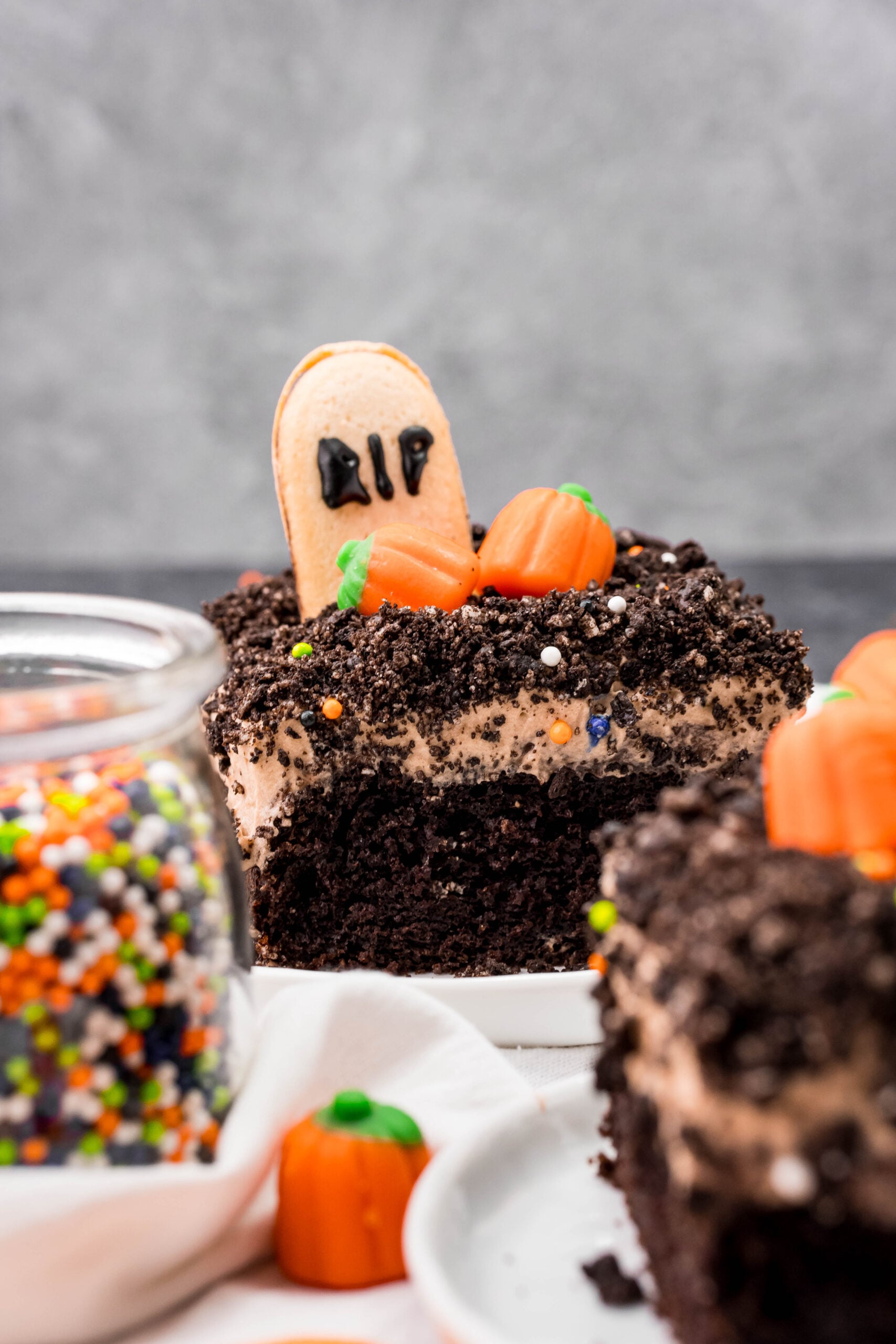 Halloween Dirt Cake slice on a white plate with aglass jar of halloween colored sprinkles next to the slice