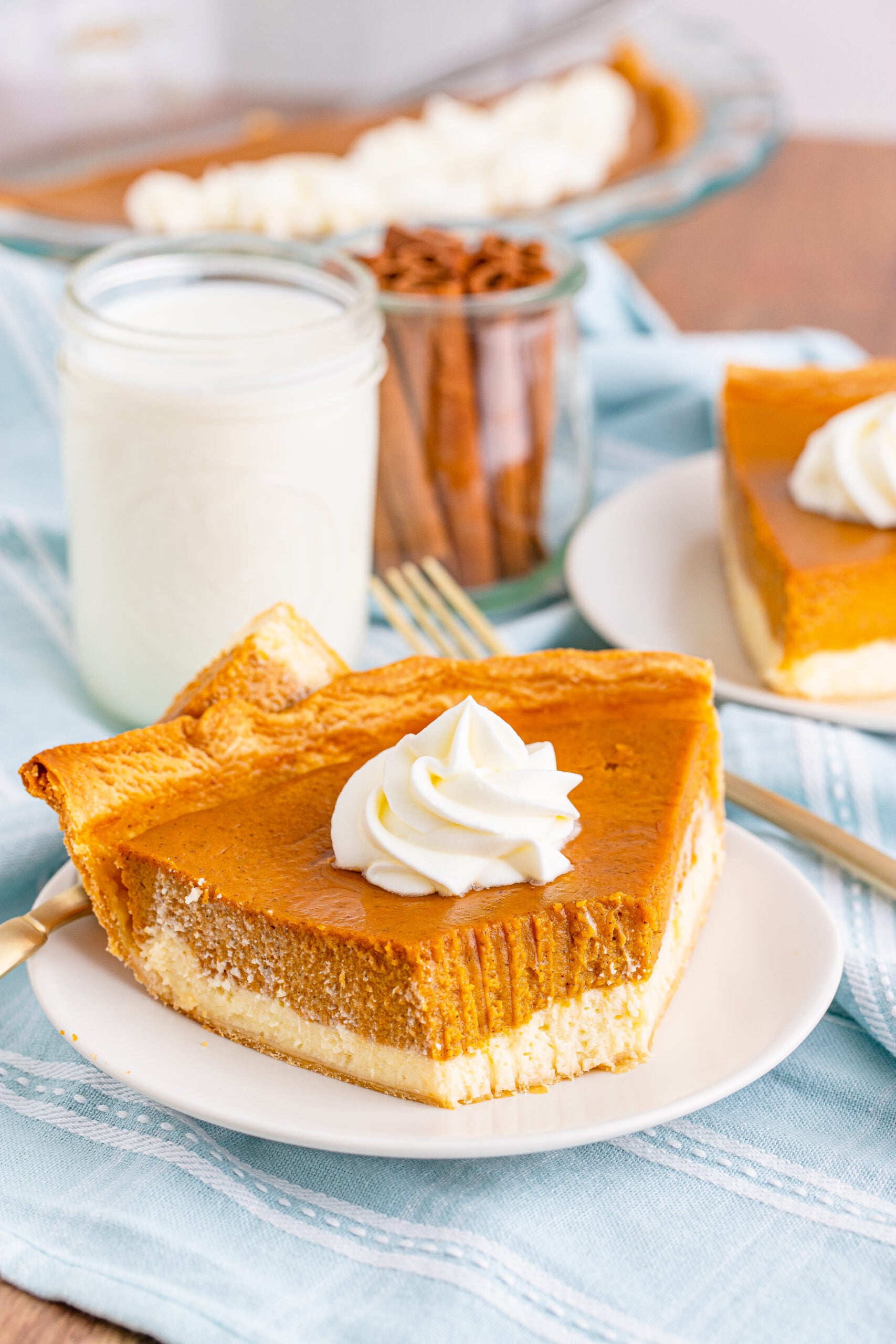 Pumpkin Pie Cheesecake slice on a white plate with a glass of milk and a glass filled with cinnamon sticks behind it