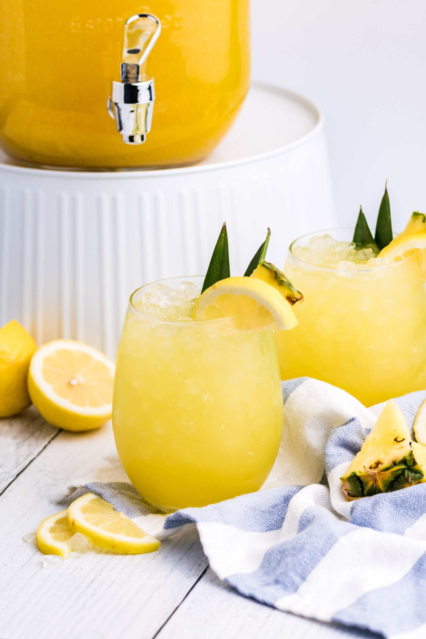 Pineapple Lemonade in a clear glass with ice on a white wood table with a punch jar in the background
