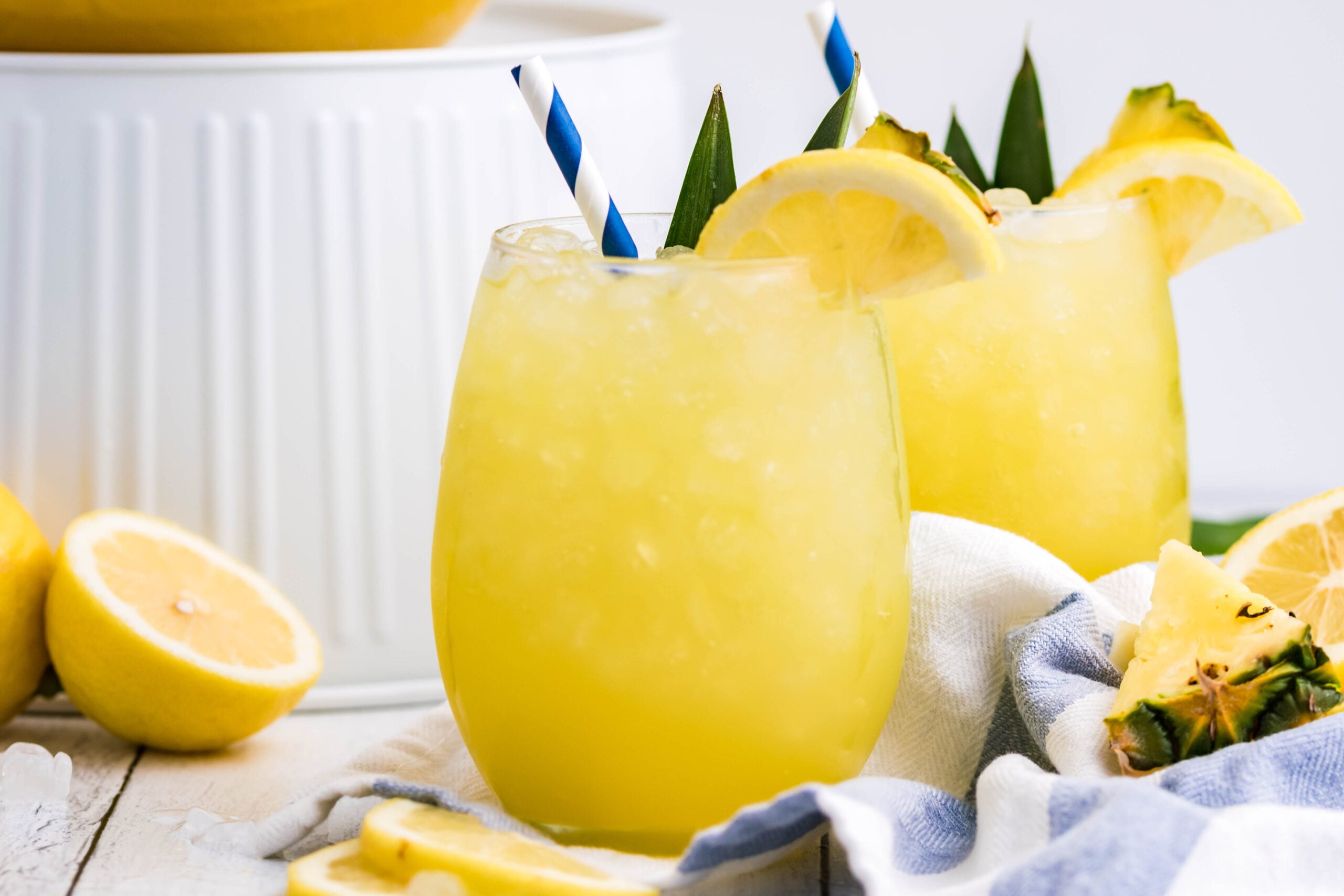Pineapple Lemonade in a clear glass with ice, a blue striped straw and sliced lemon
