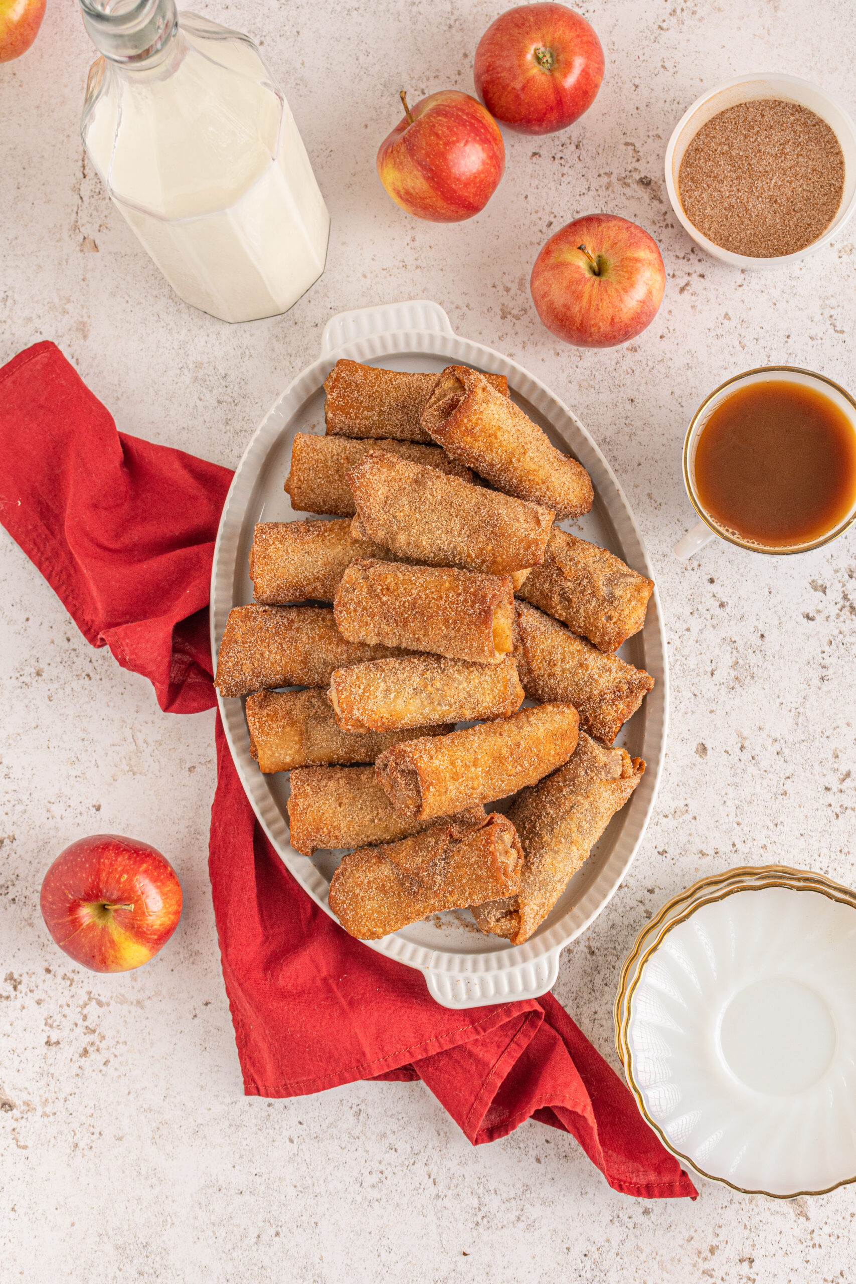 Overhead view of egg rolls stacked on a white platter with apples and a red linen in the background.