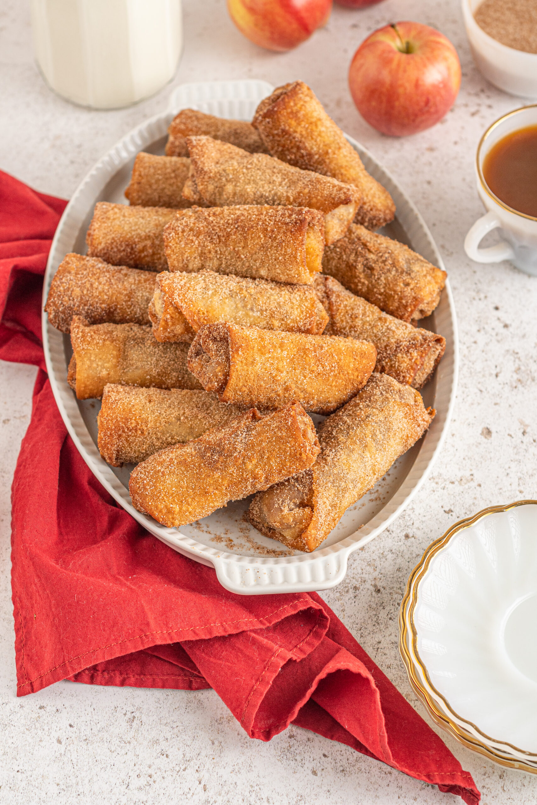 Egg rolls stacked on a white platter with apples and a red linen in the background.