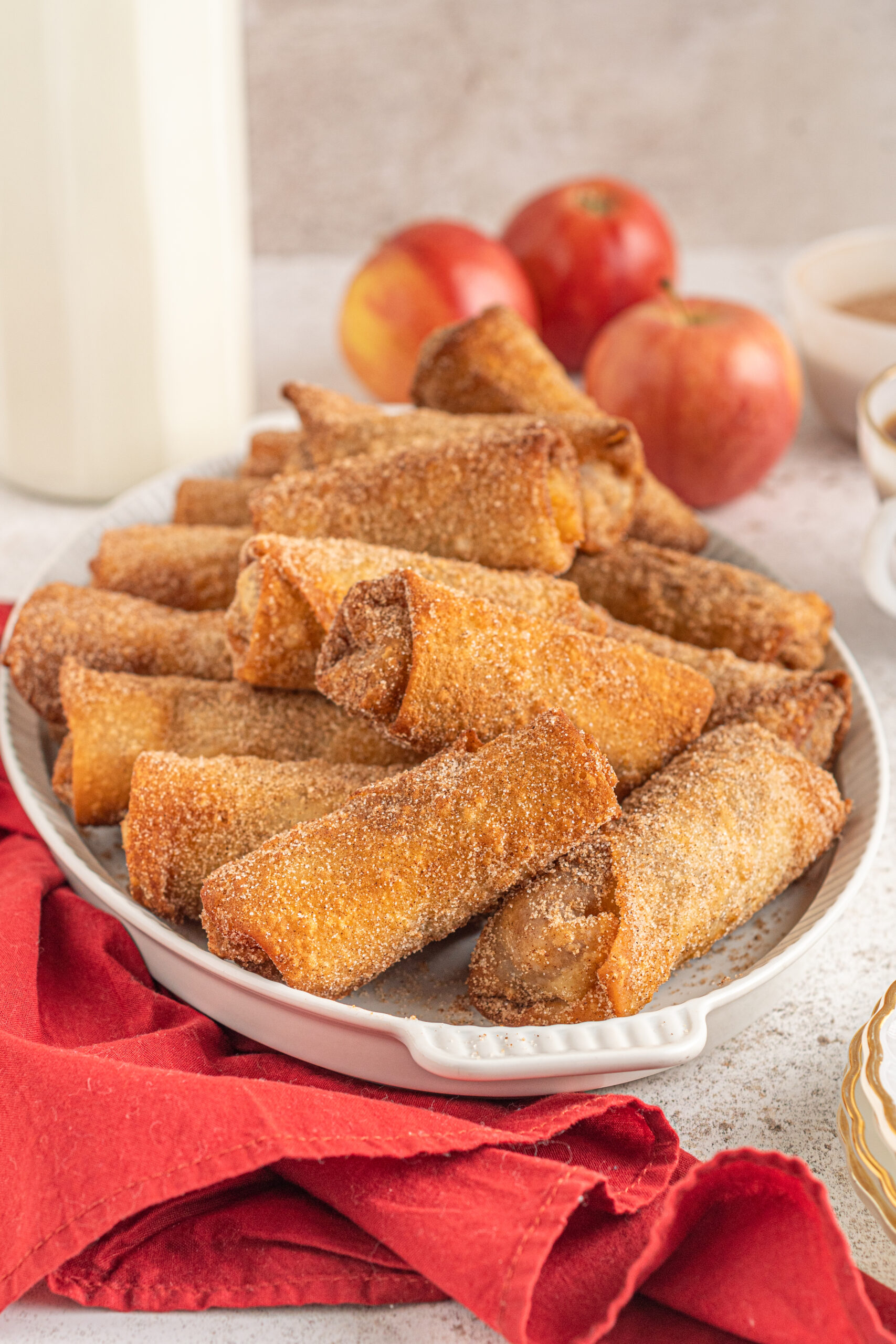 Apple pie egg rolls stacked on a white platter with apples and a red linen in the background.