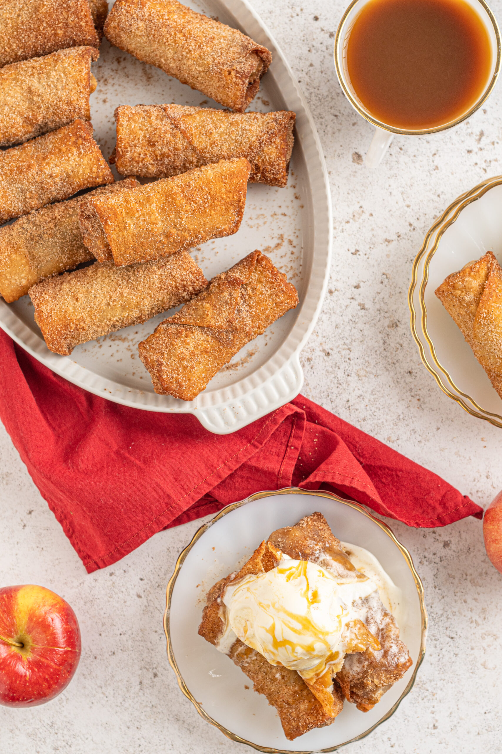 Overhead view of Apple pie egg rolls stacked on a white platter with apples and a red linen in the background.