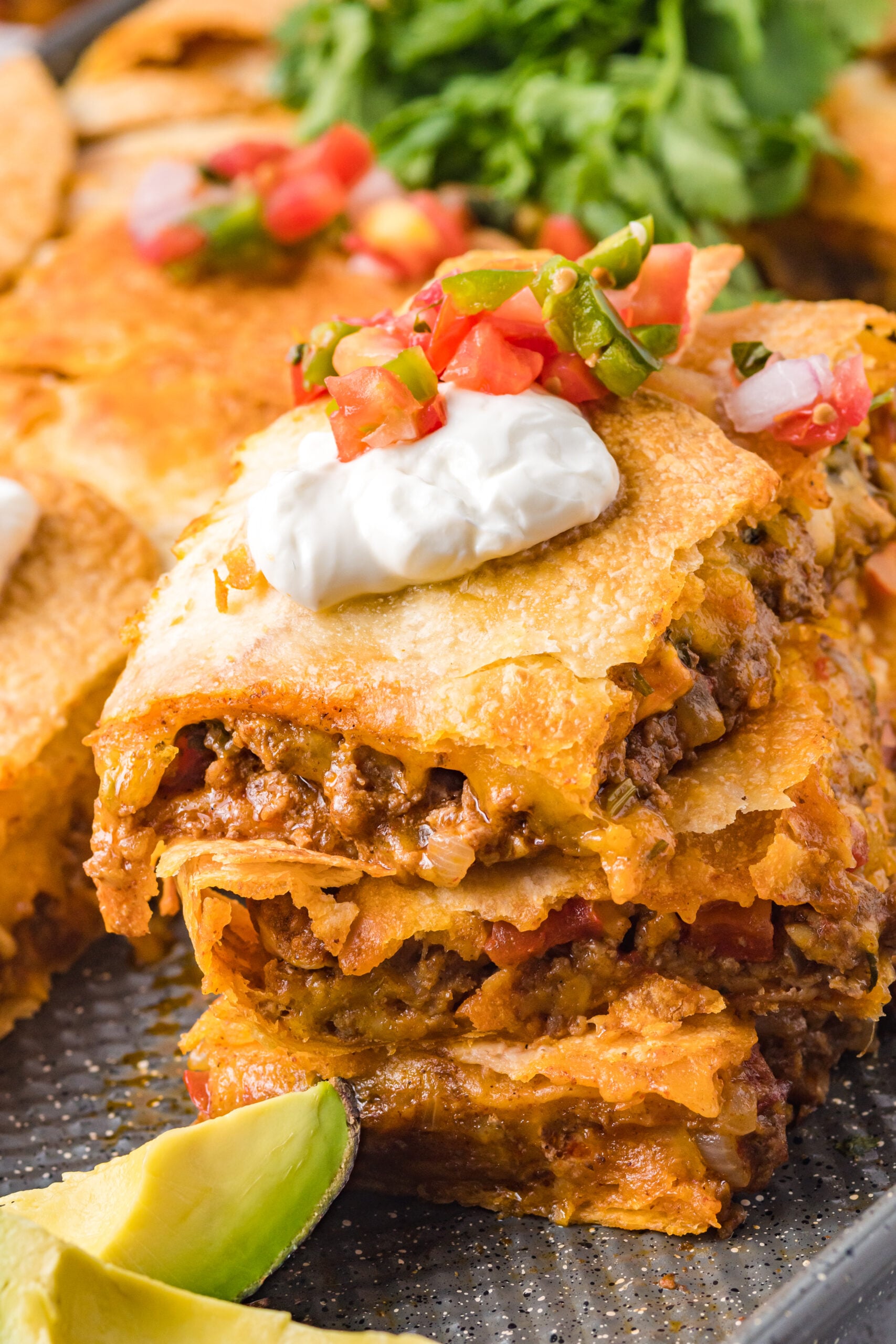 Close up of three slices of sheet pan tacos stacked on the baking sheet topped with sour cream and Pico de Gallo 