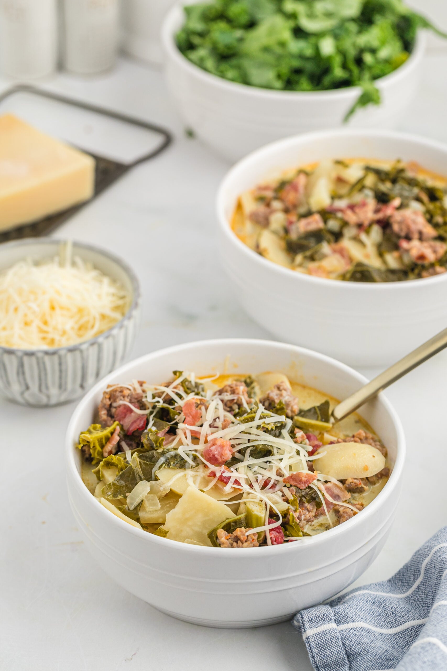45 degree angle view of the Toscana soup in a white bowl topped with shredded cheese. Shredded cheese and kale in a white bowl in the background. 