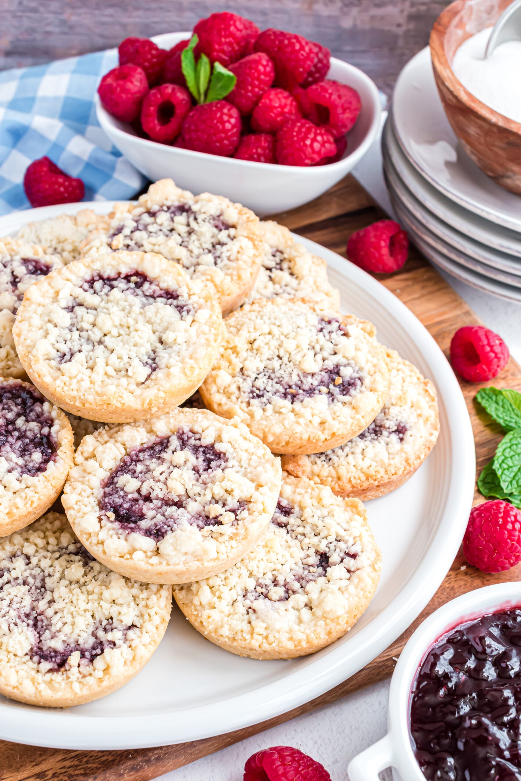 Raspberry Cookies stacked on a white plate on a wooden cutting board with fresh raspberries in the background.