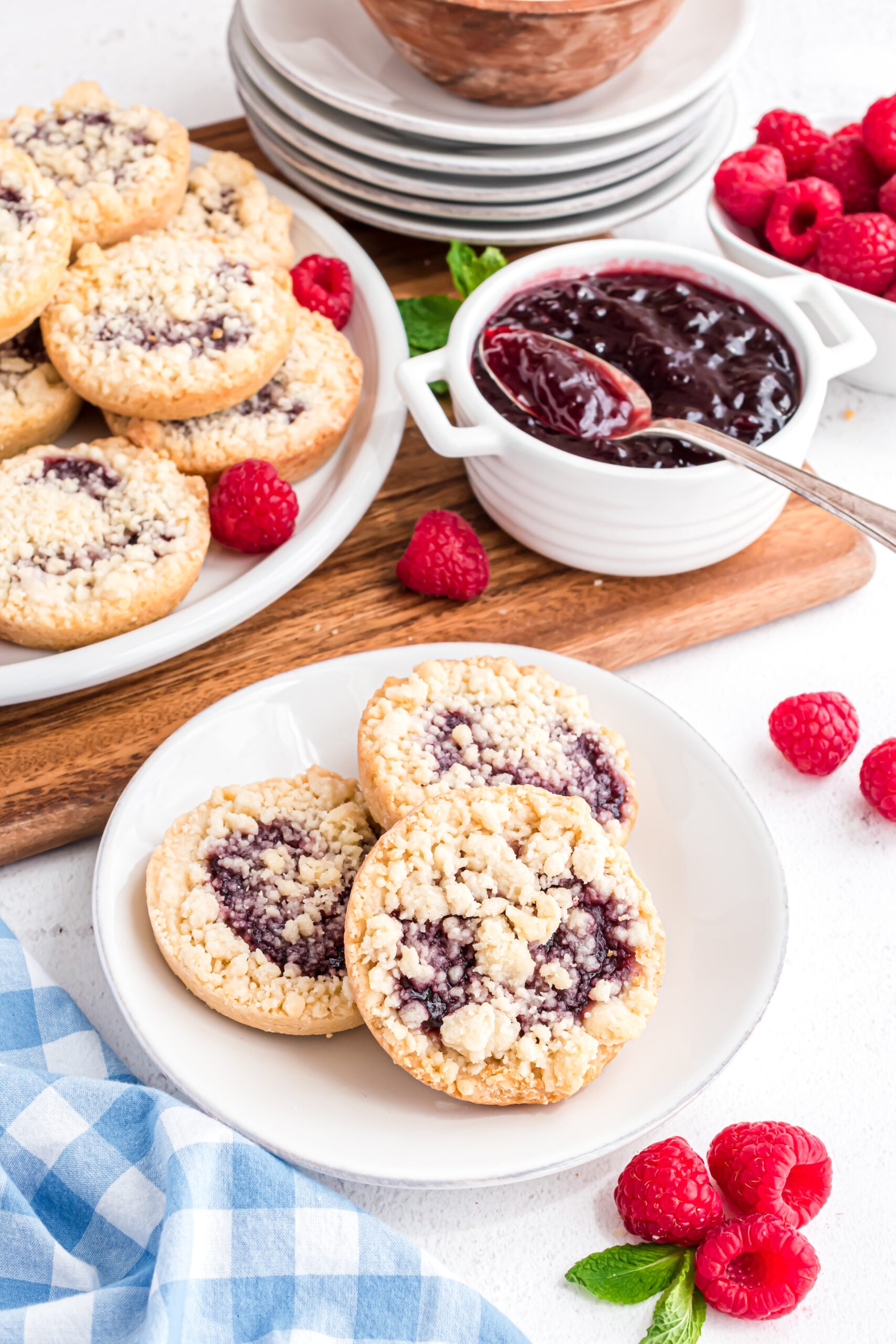 Three cookies on a white plate with a blue and white linen and fresh raspberries in the background.
