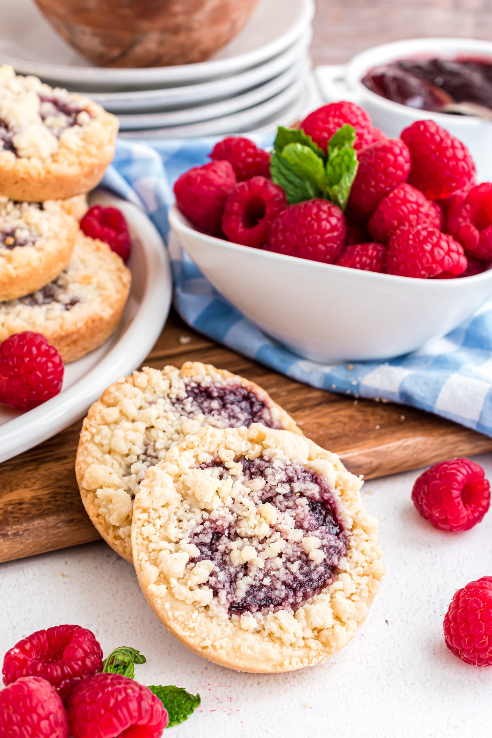 Two raspberry cookies propped against a wooden cutting board with fresh raspberries in a white bowl.