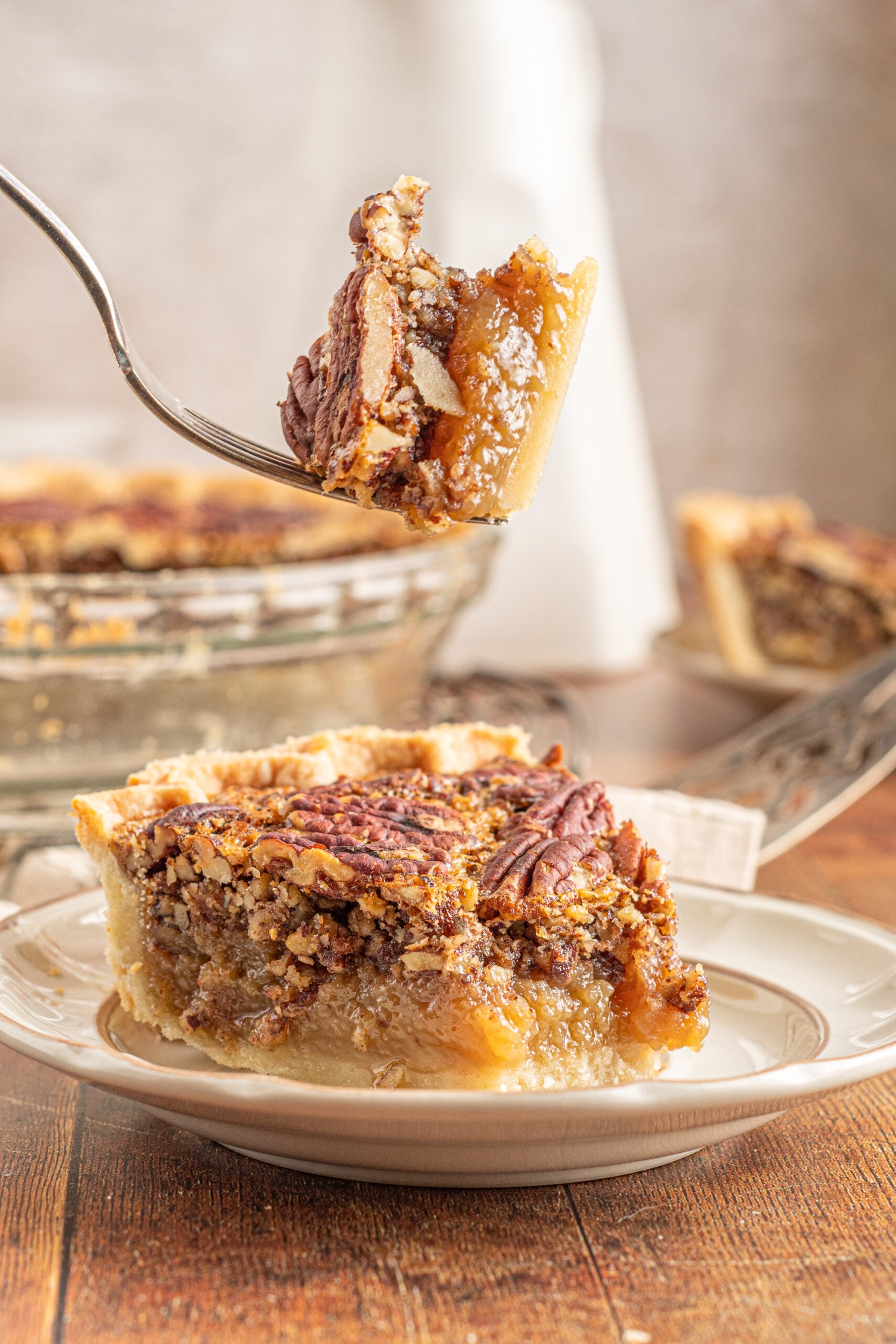 A piece of pecan pie on a tan and brown plate with a bite of pie being removed with a silver fork.