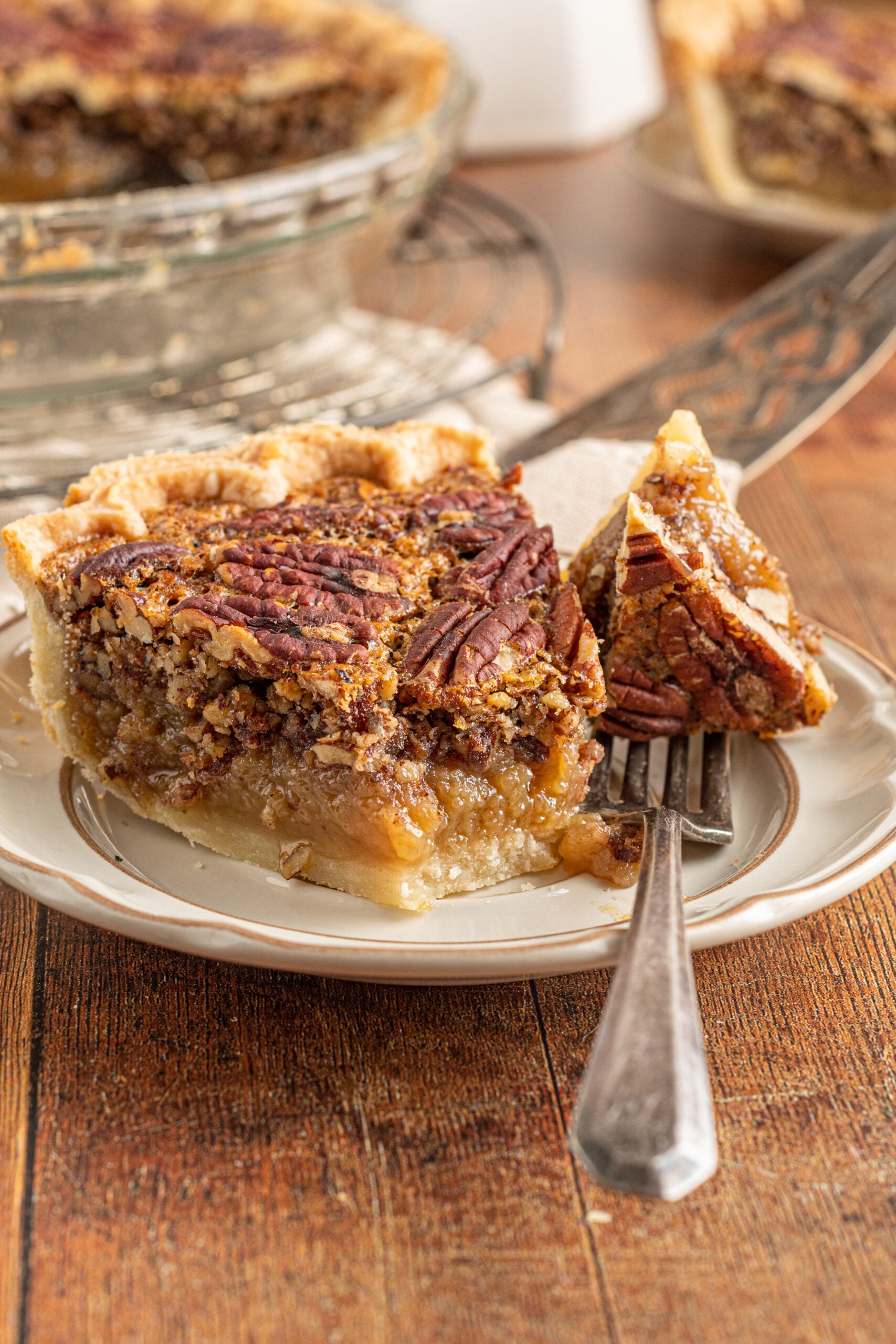 A piece of pecan pie on a tan and brown plate with a bite of pie being removed with a silver fork.