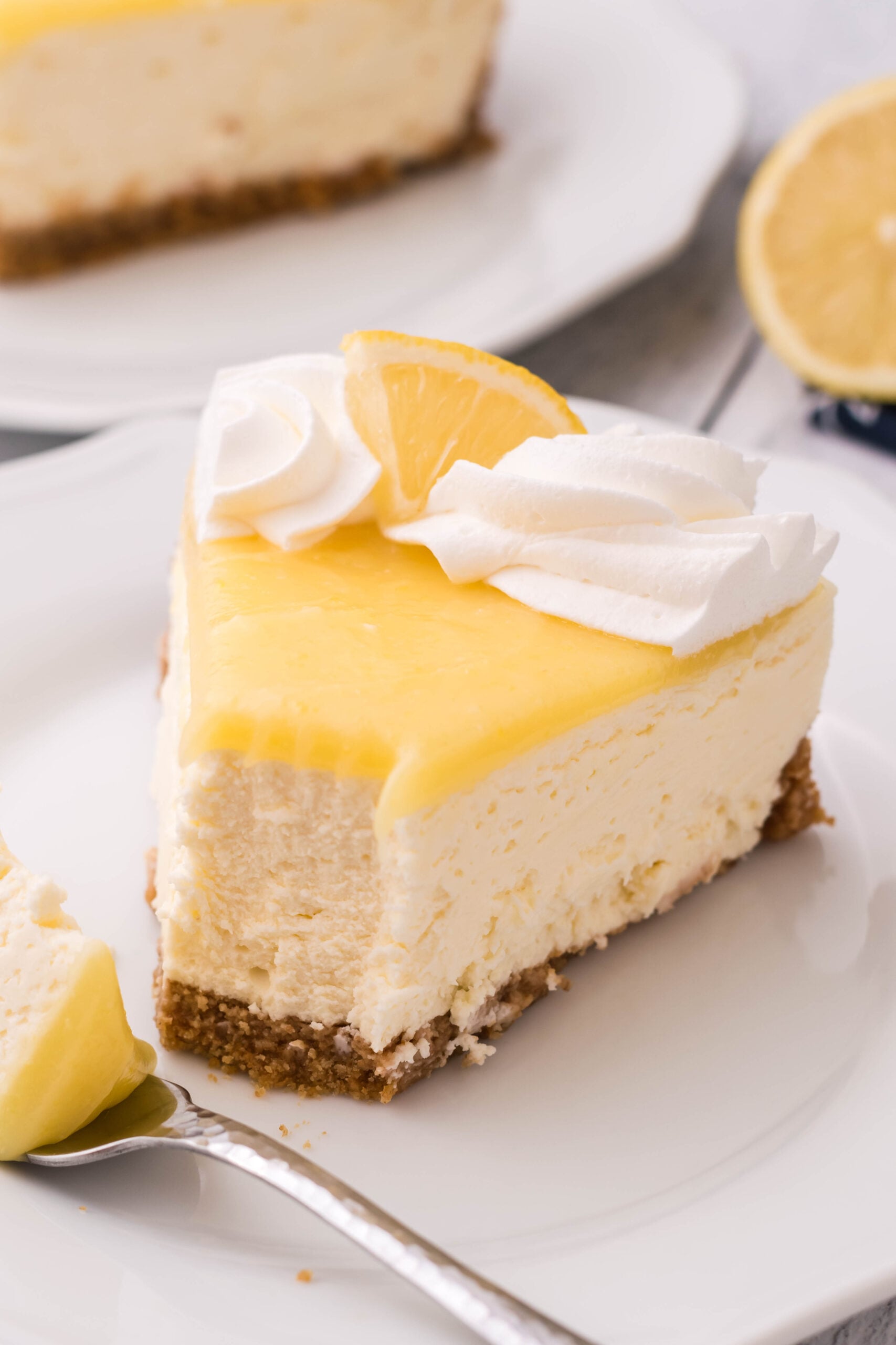 A slice of cheesecake topped with whipped cream and a lemon slice on a white plate with a bite removed.