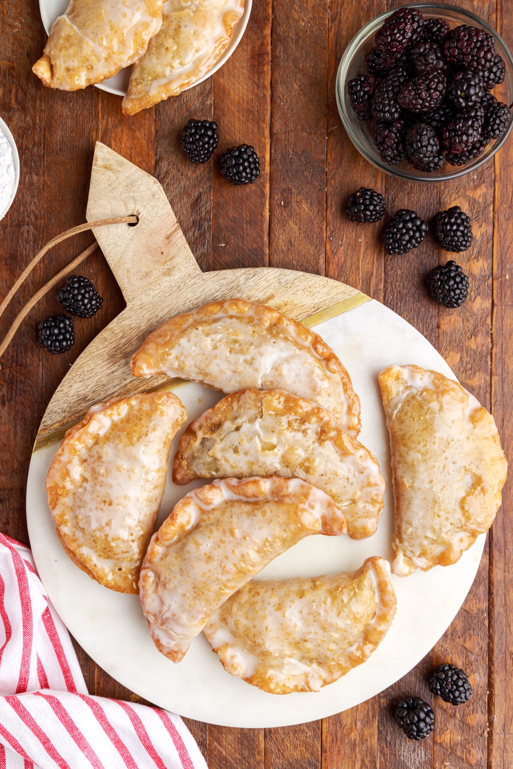 Overhead shot of blackberry hand pies stacked on a marble serving tray with fresh blackberries on a dark wood background. 