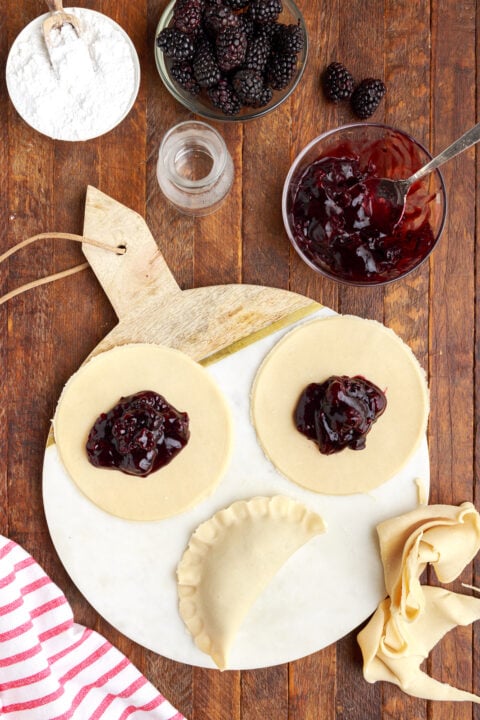 three circle of pie dough on a marble serving tray with blackberry jam in the center of each circle. One is folded in half showing the correct way to fold a hand pie. 