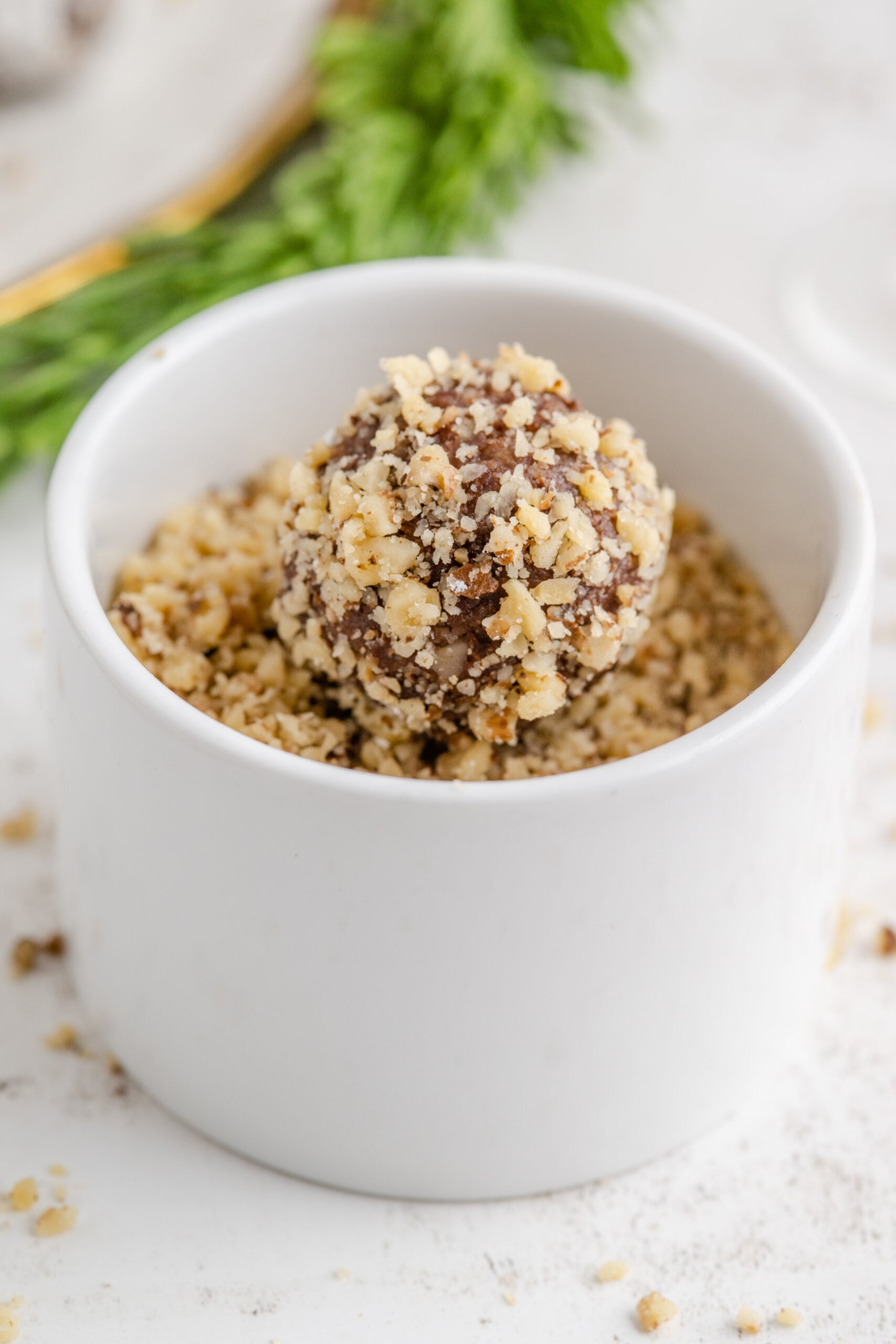 A rum ball covered in crushed nuts in a small white bowl of crushed nuts.