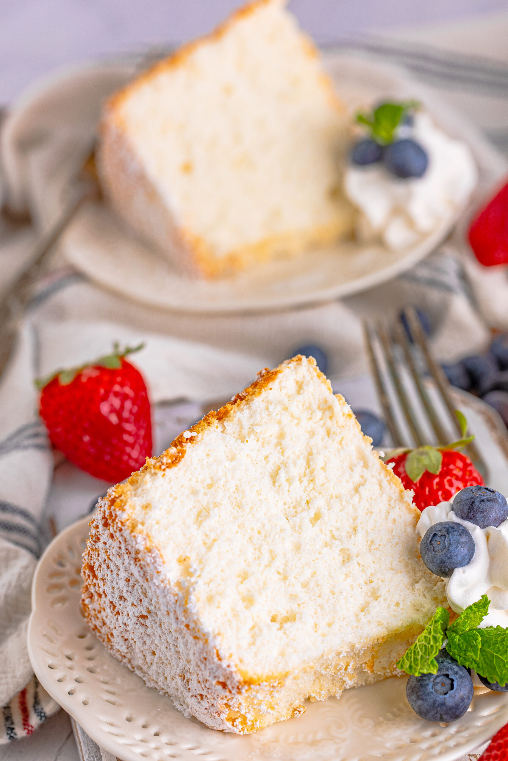 A piece of Angel Food Cake on a white plate with whipped cream, fresh berries, and mint. 