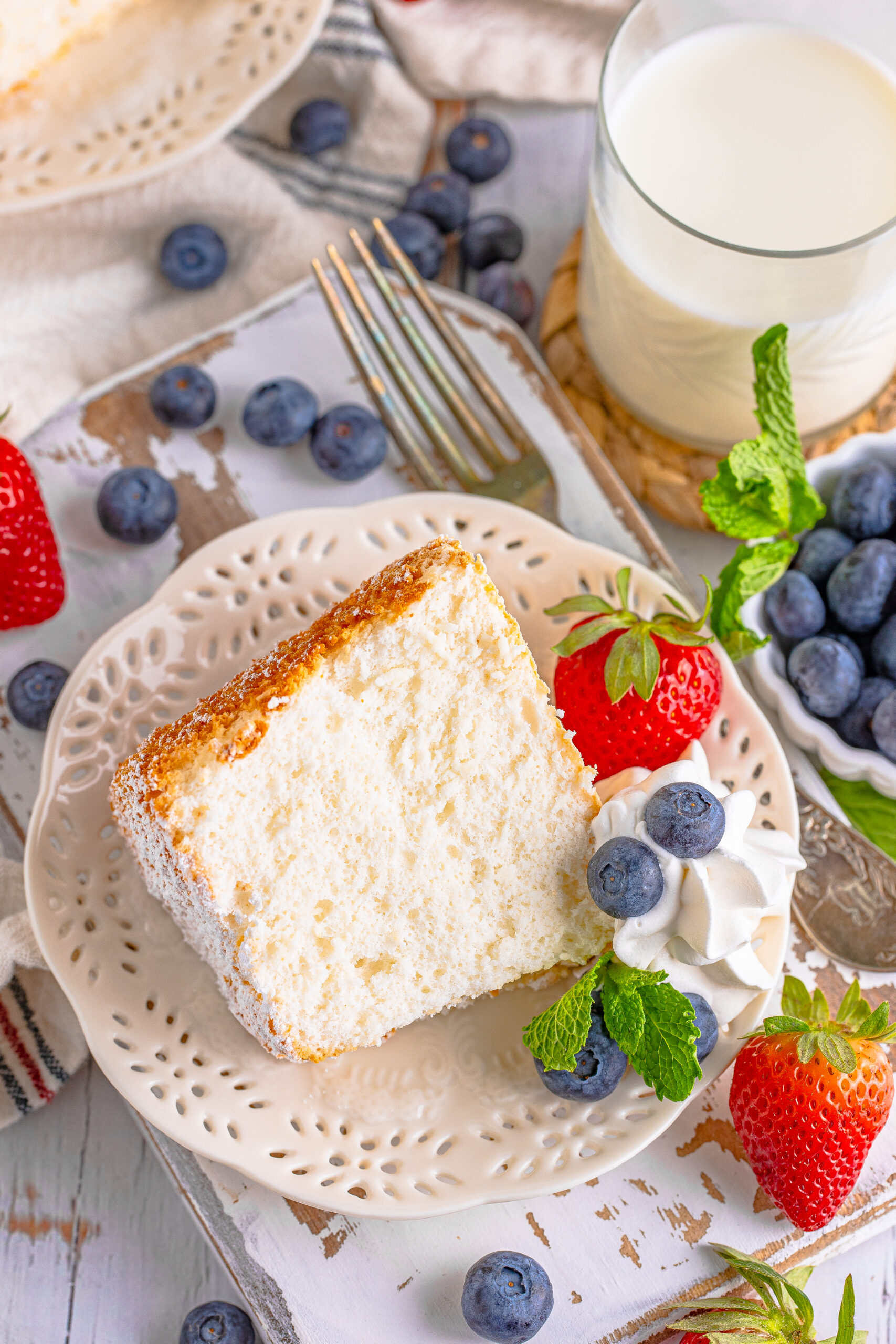 A piece of Angel Food Cake on a white plate with whipped cream, fresh berries, and mint. 