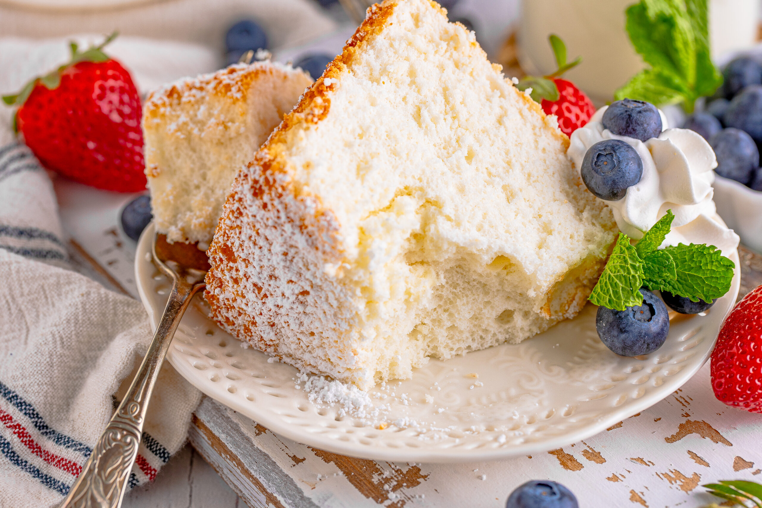 A piece of Angel Food Cake on a white plate with a bite removed on a silver fork with whipped cream, fresh berries, and mint